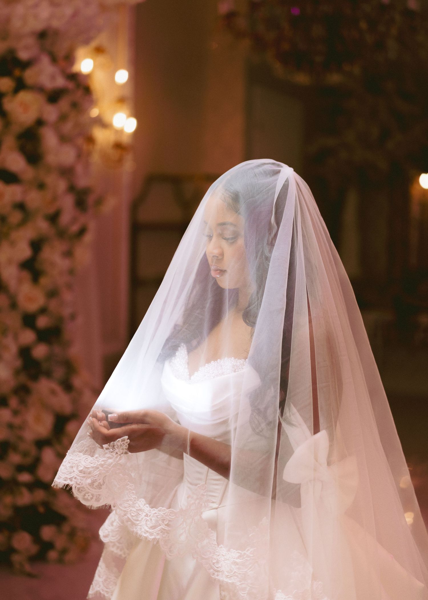 Bride in white lace wedding dress and flowing veil, standing among pink floral arrangements in soft romantic lighting.