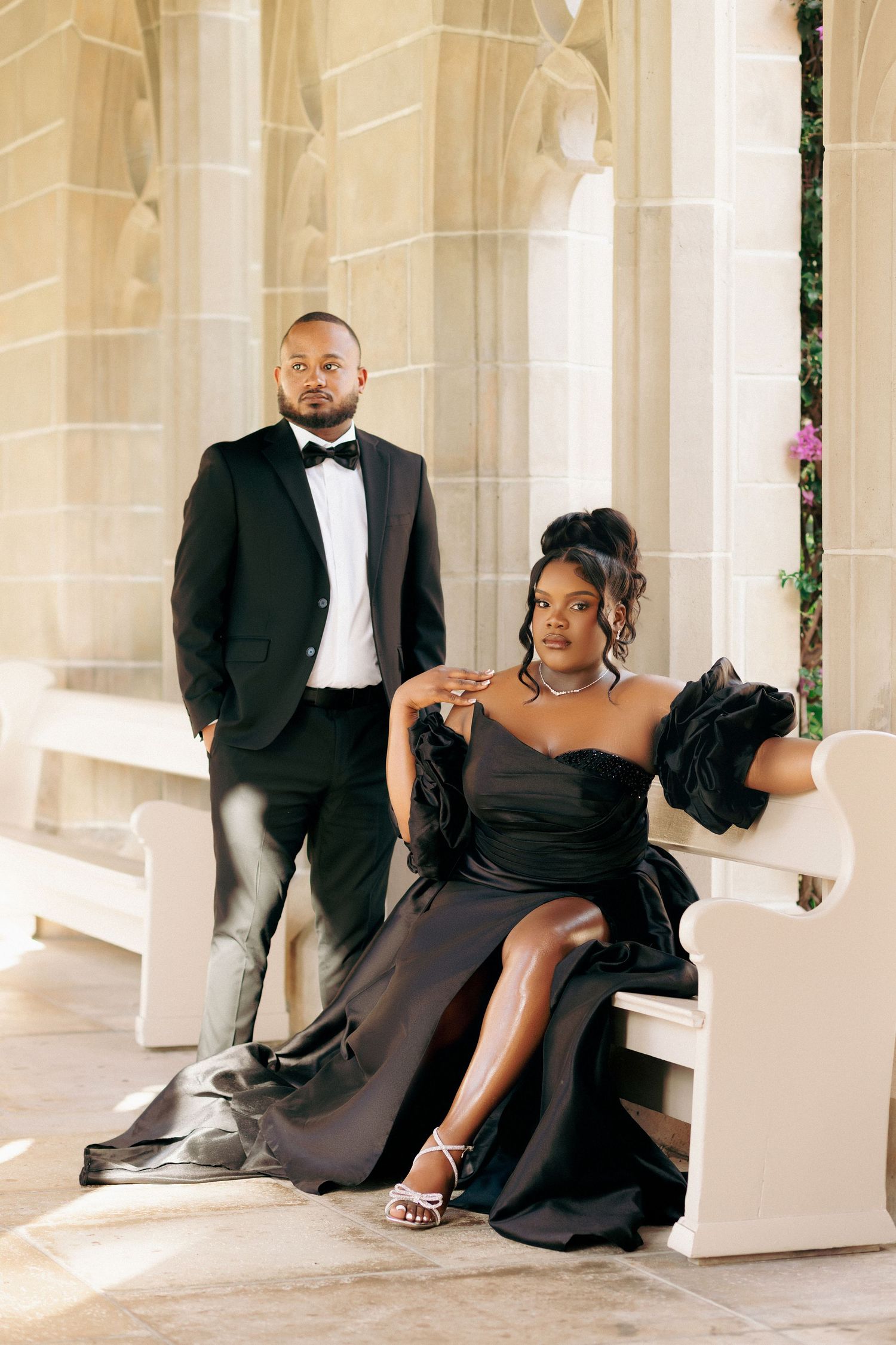 Elegant couple in matching black formal attire posing in a sunlit stone archway, man standing, woman seated on white bench.