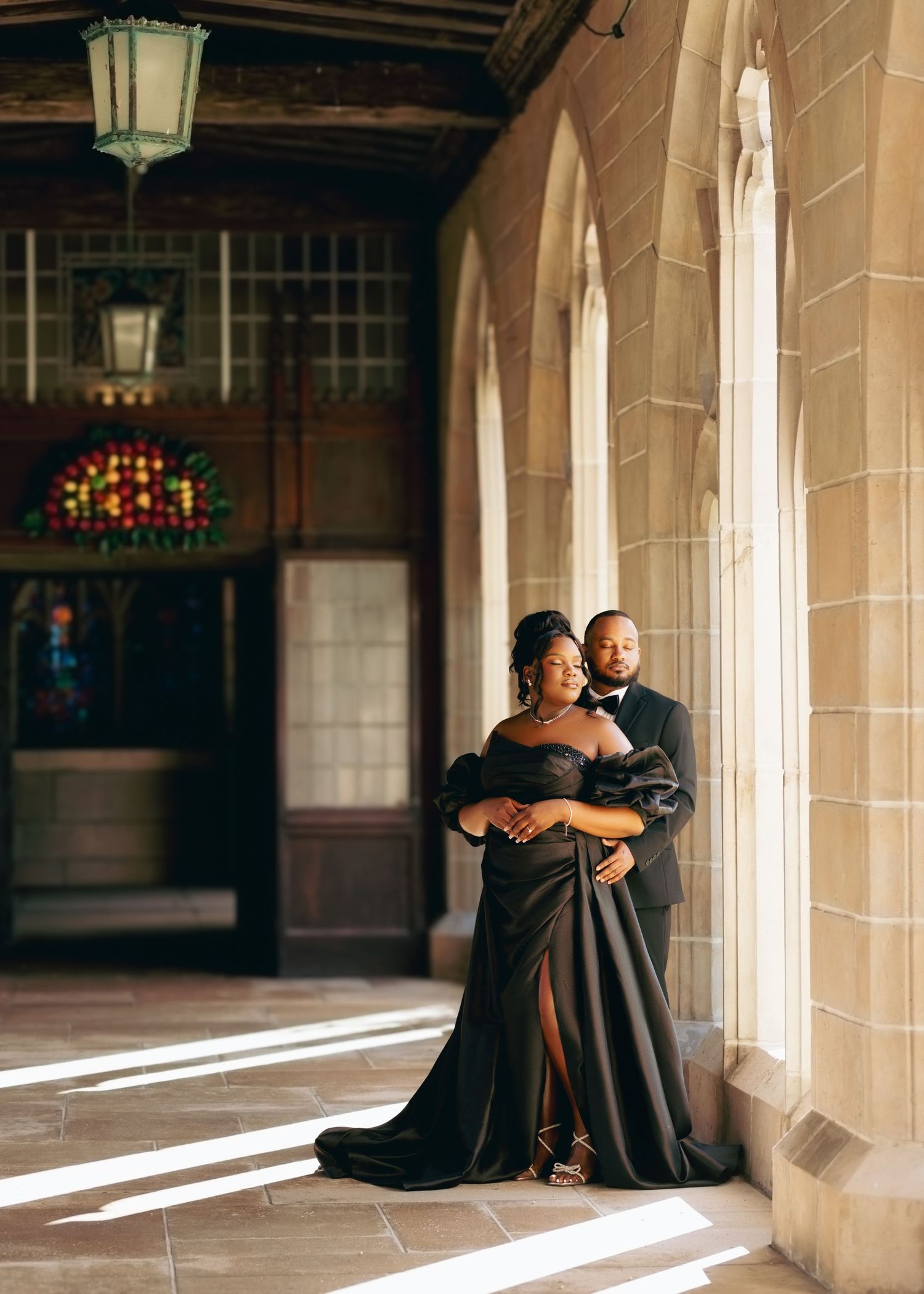 Elegant couple in formal attire embrace in a sunlit stone archway corridor with vintage architecture and warm lighting.