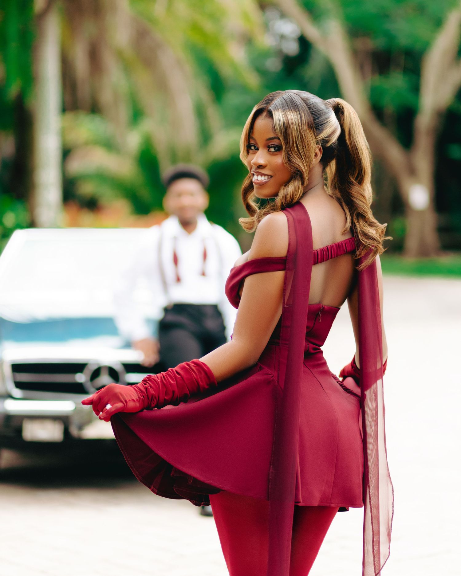 A person in a burgundy formal outfit with tulle details poses elegantly by a car in an outdoor setting.
