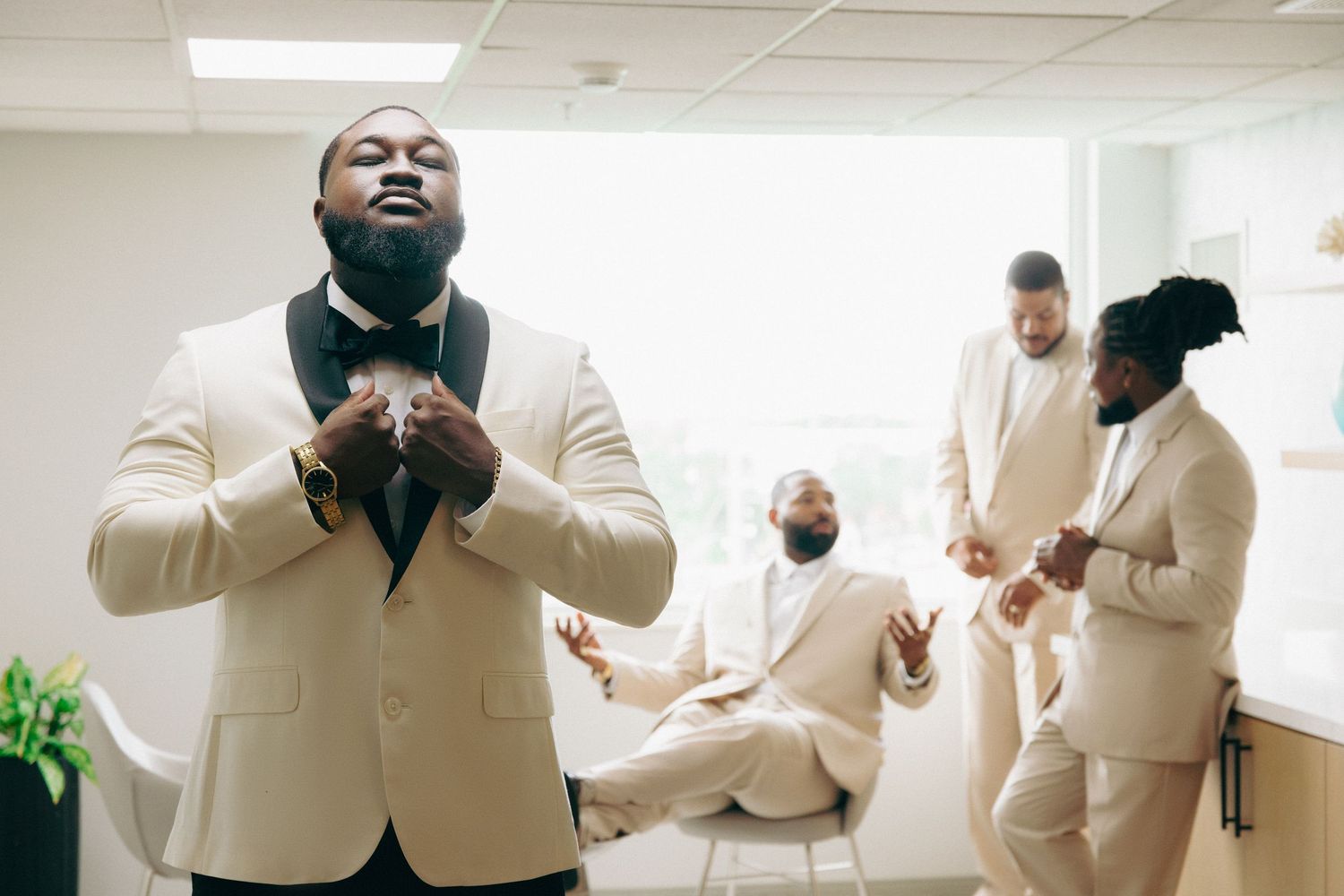 Groomsmen in cream colored suits getting ready in a bright room before a wedding ceremony.