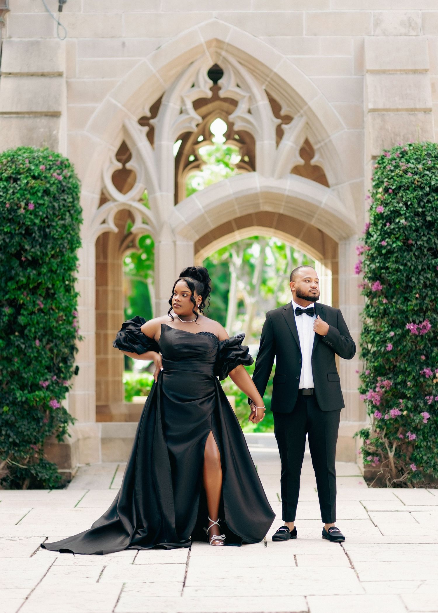 Elegant couple in matching black formal wear posing before Gothic stone archway surrounded by lush greenery.