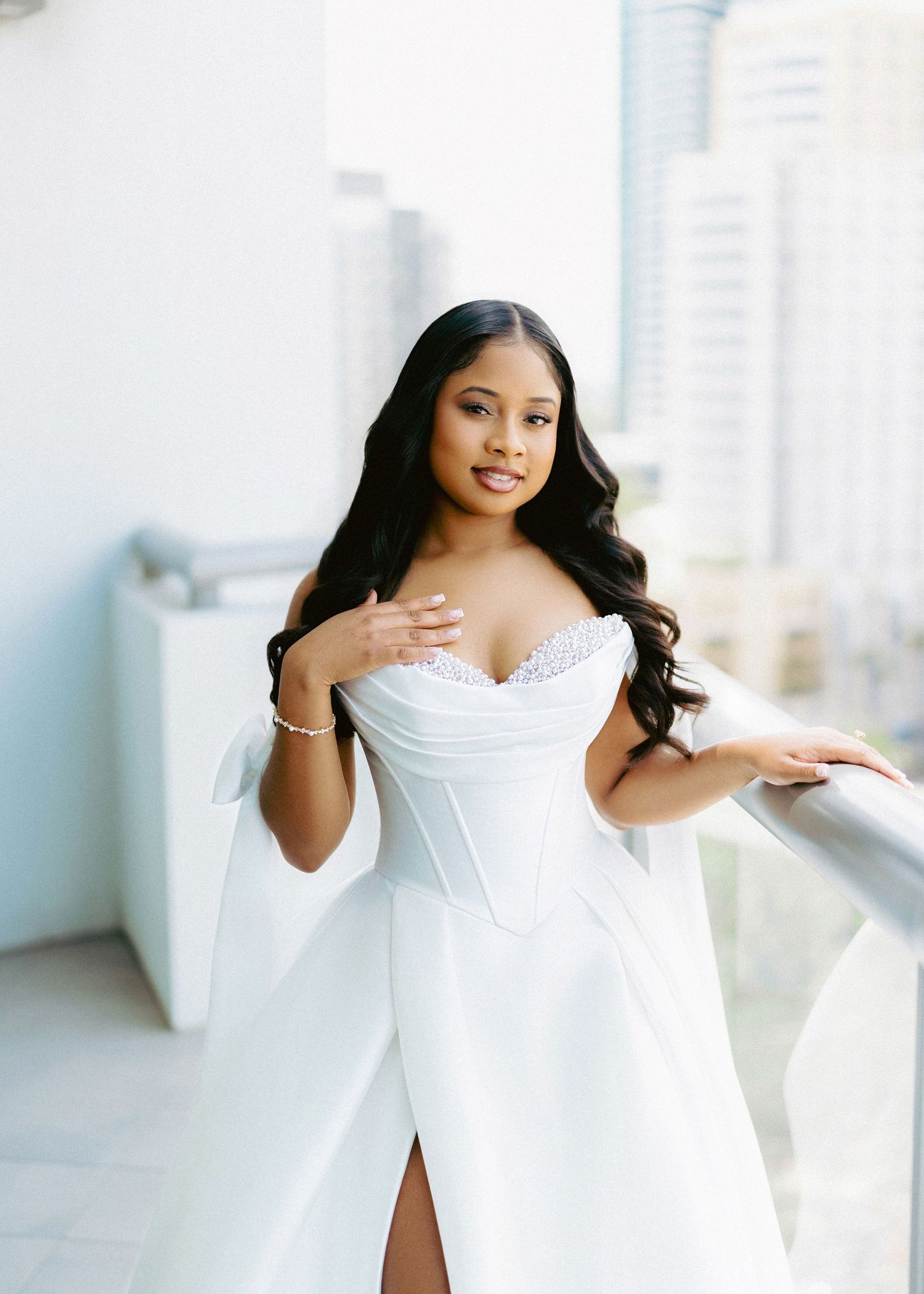 Elegant bride in white off-shoulder corset gown with cape sleeves poses on a balcony with city views.
