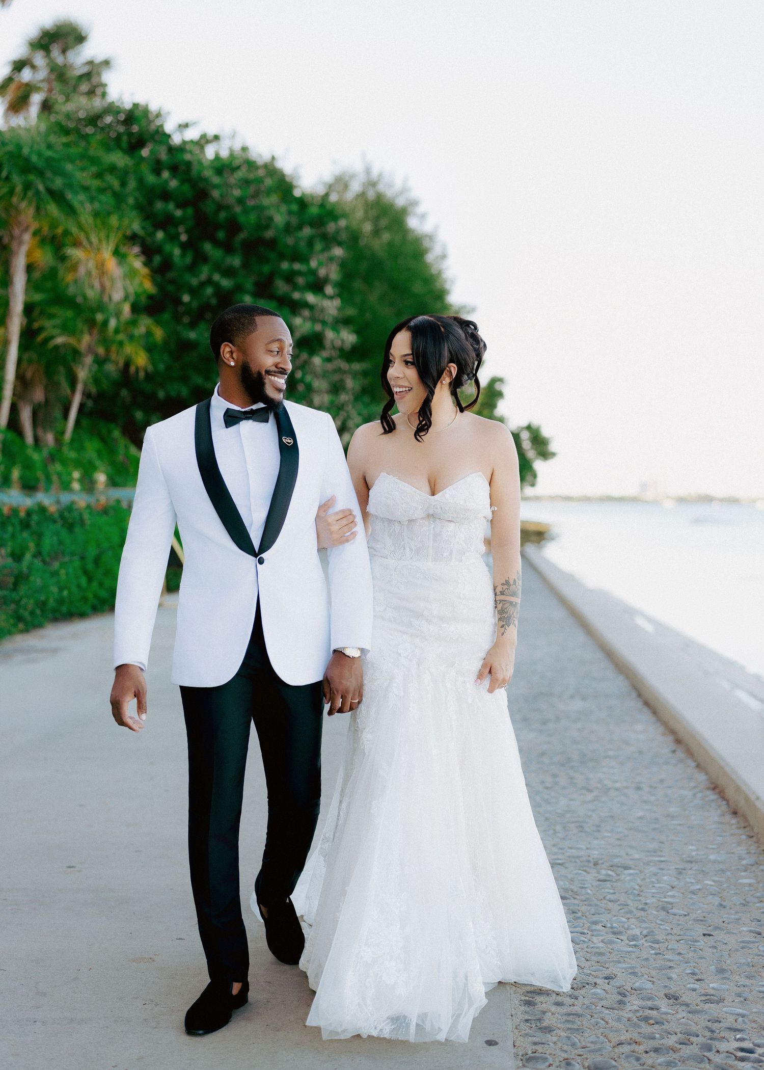 Bride and groom walking hand in hand along waterfront path, smiling at each other on wedding day.