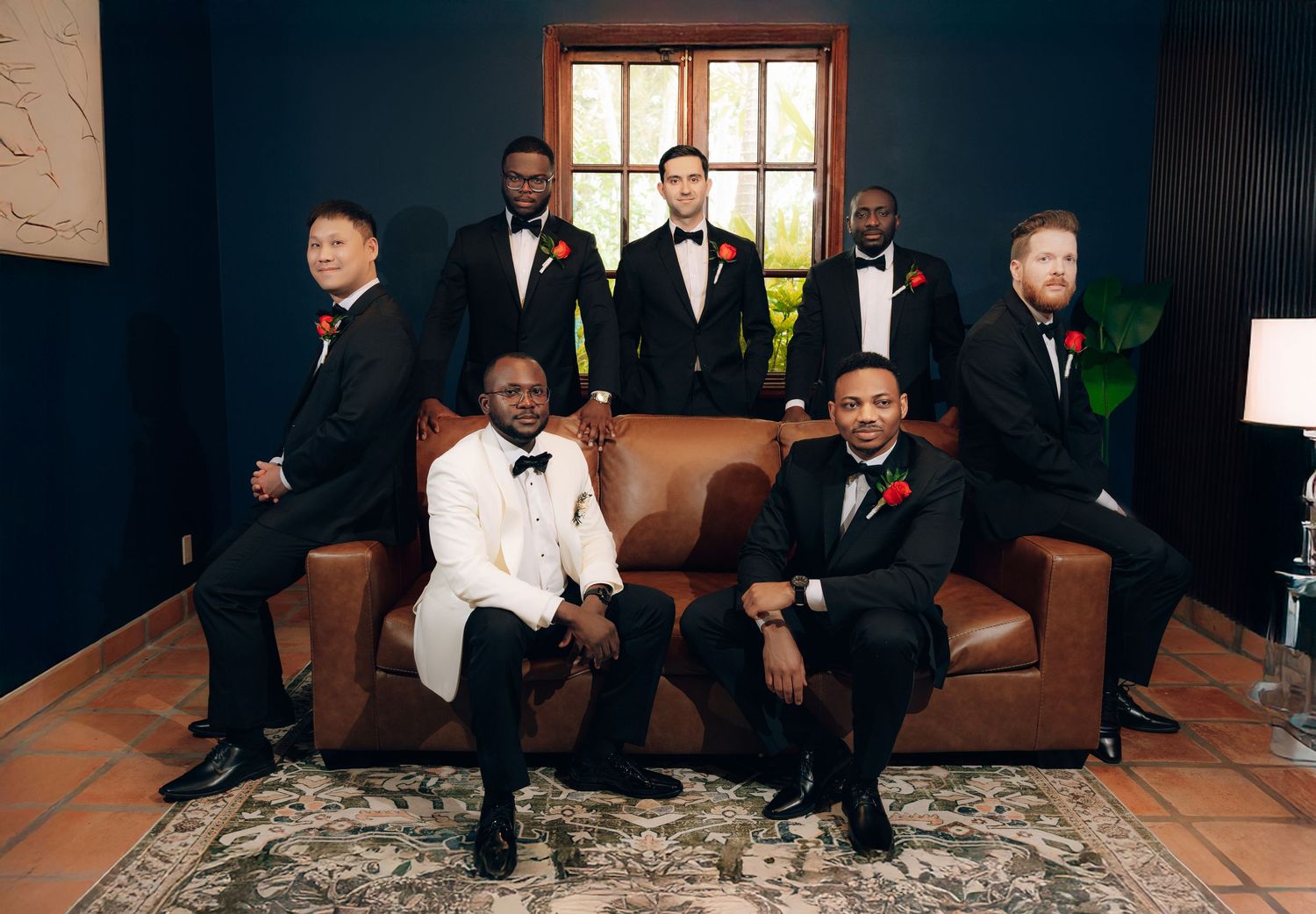 Wedding party in formal attire poses together on elegant furniture against dark backdrop.