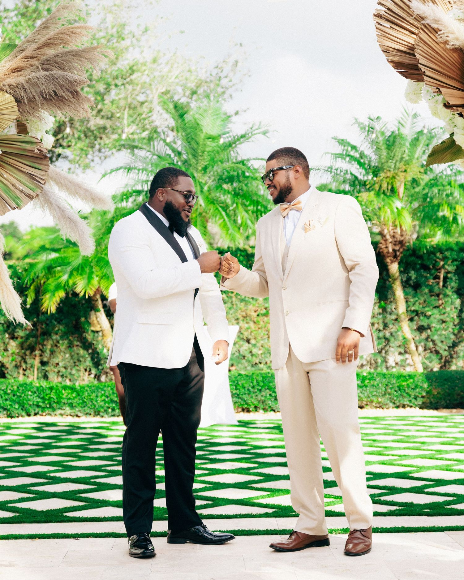Two men in formal white suits share a moment in a tropical garden setting.