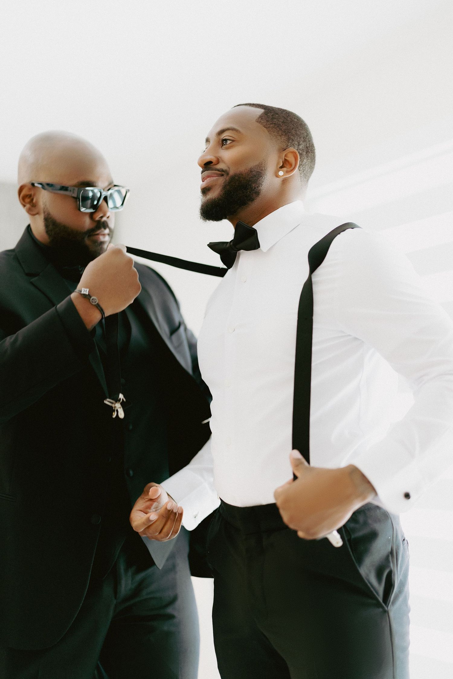 Two men in formal attire sharing a moment, one adjusting the other's bow tie indoors.