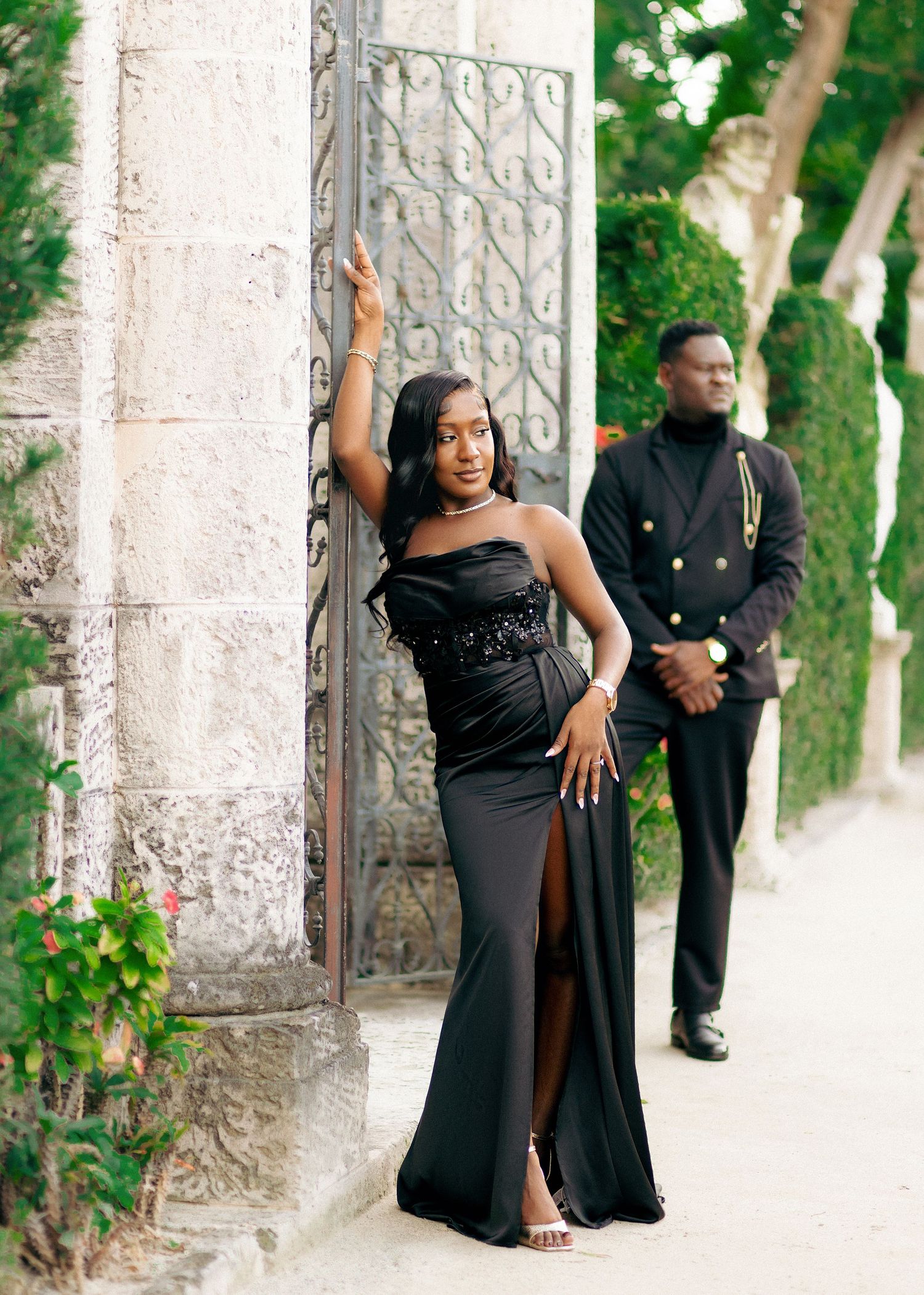 A group in formal black attire poses against stone pillars in an outdoor garden setting.