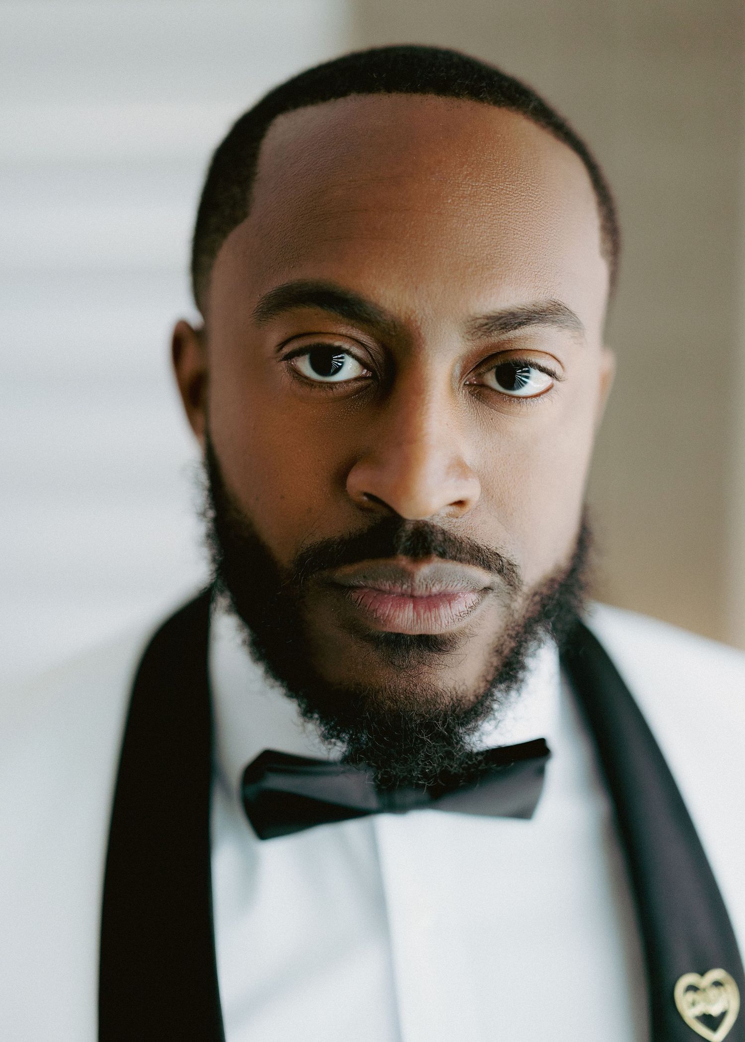 Close-up portrait of groom in white tuxedo with black bow tie and gold heart lapel pin.