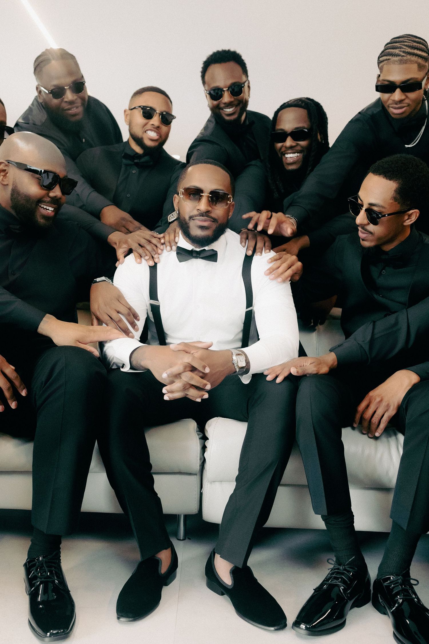 A well-dressed man in white shirt and bow tie sits on a sofa surrounded by eight men in black outfits and sunglasses.