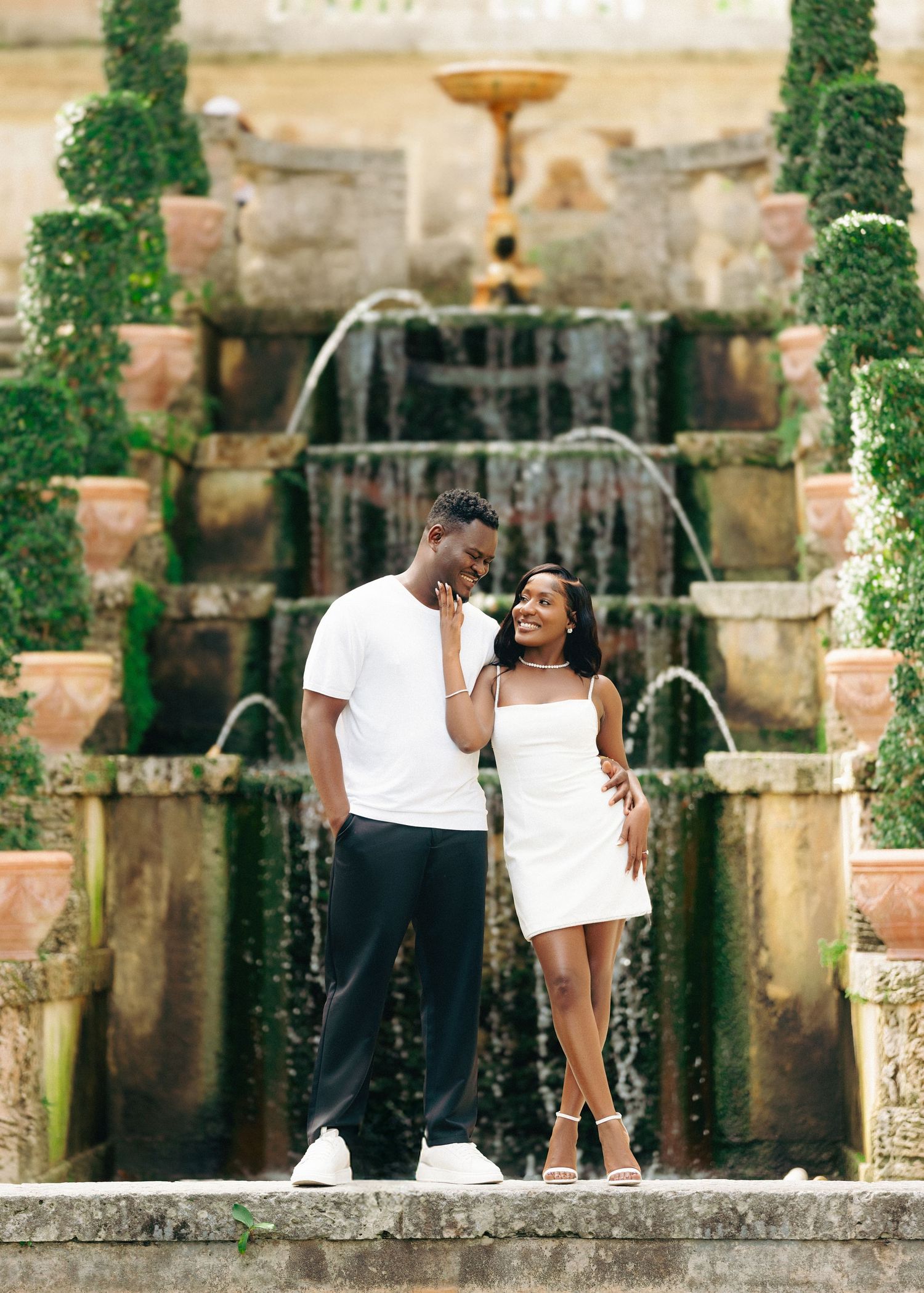 A couple shares a romantic moment in front of a stone fountain in an outdoor garden setting.