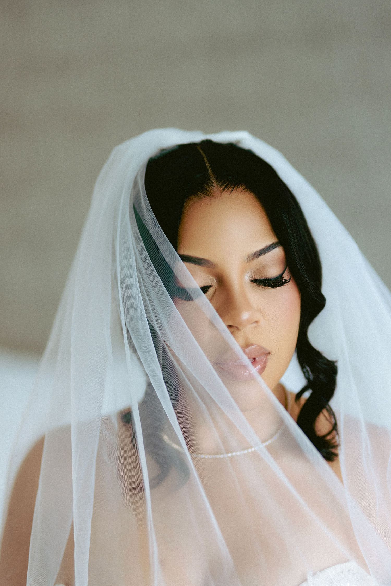 Beautiful bride wearing a flowing white veil with elegant makeup and dark curly hair.