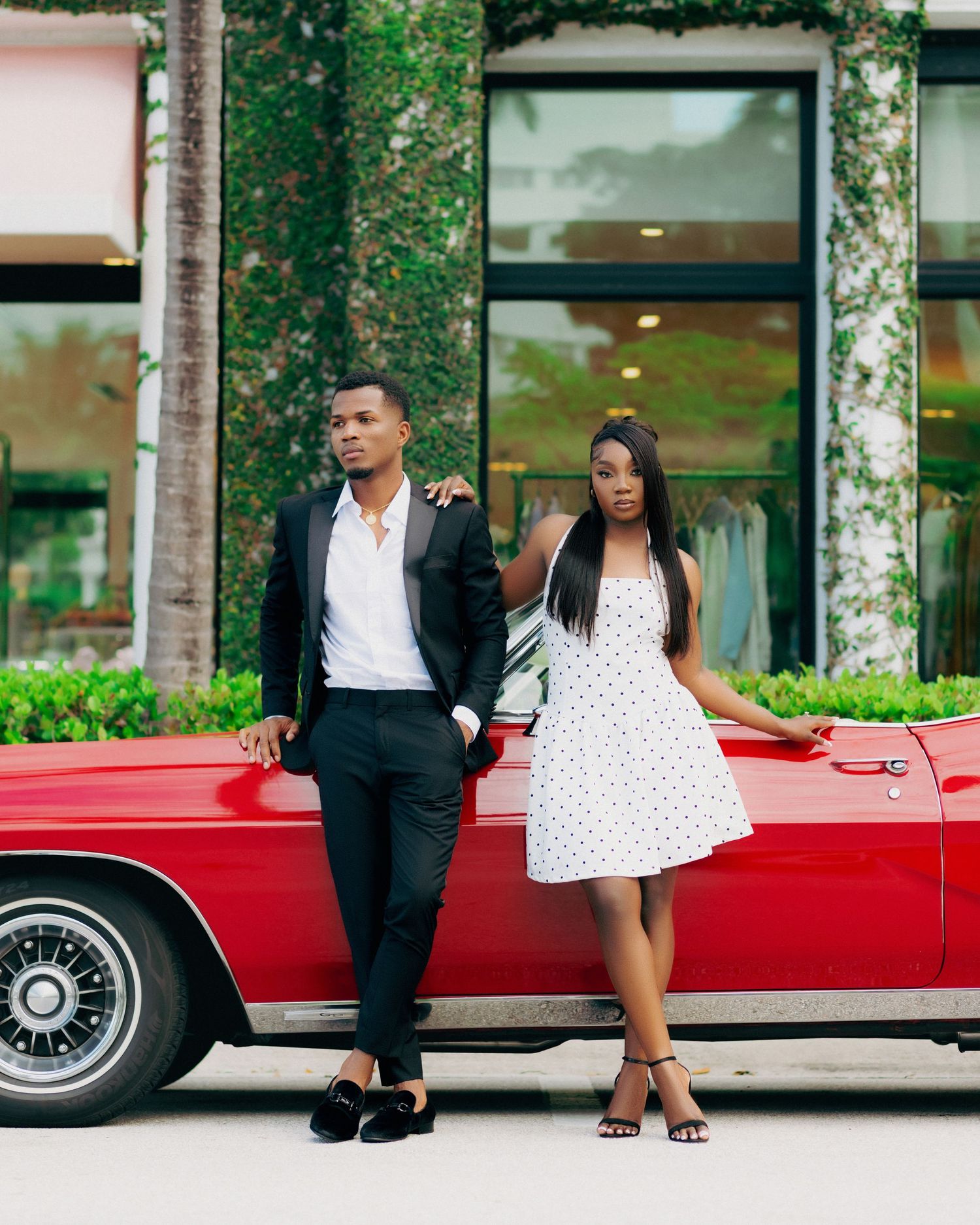 Stylish couple in formal wear sitting by a vintage red car outside a modern building.