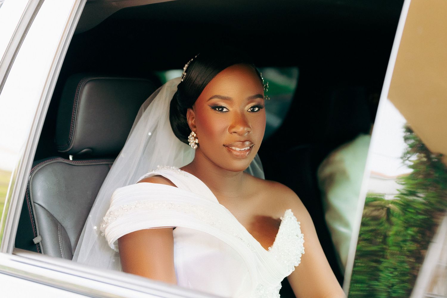 A bride wearing a white wedding dress sits elegantly in a car.