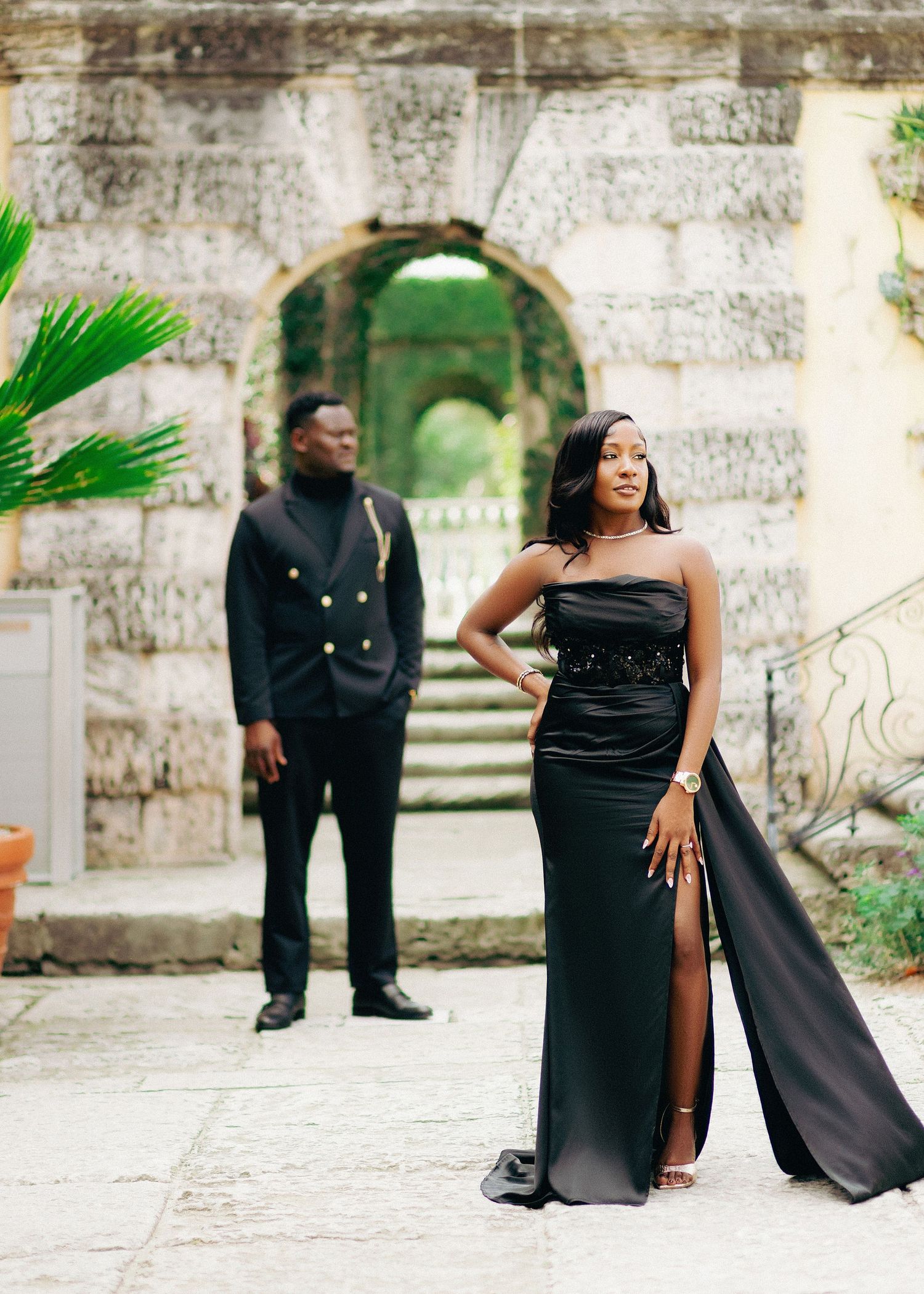 A stylish couple in formal black attire poses together at an outdoor stone venue for a photoshoot.