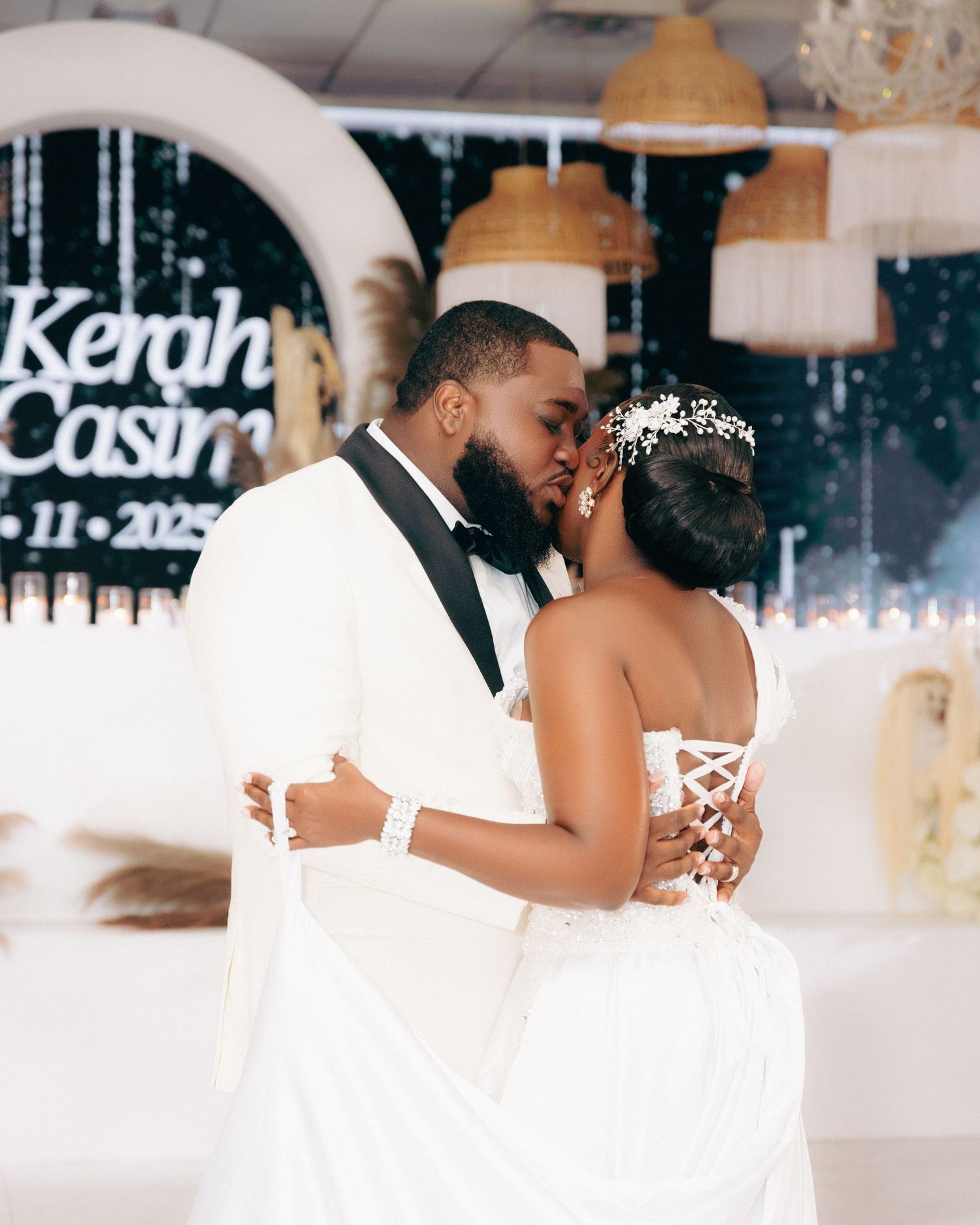 A couple in white attire share a romantic dance at their wedding reception with soft lighting and decor in the background.