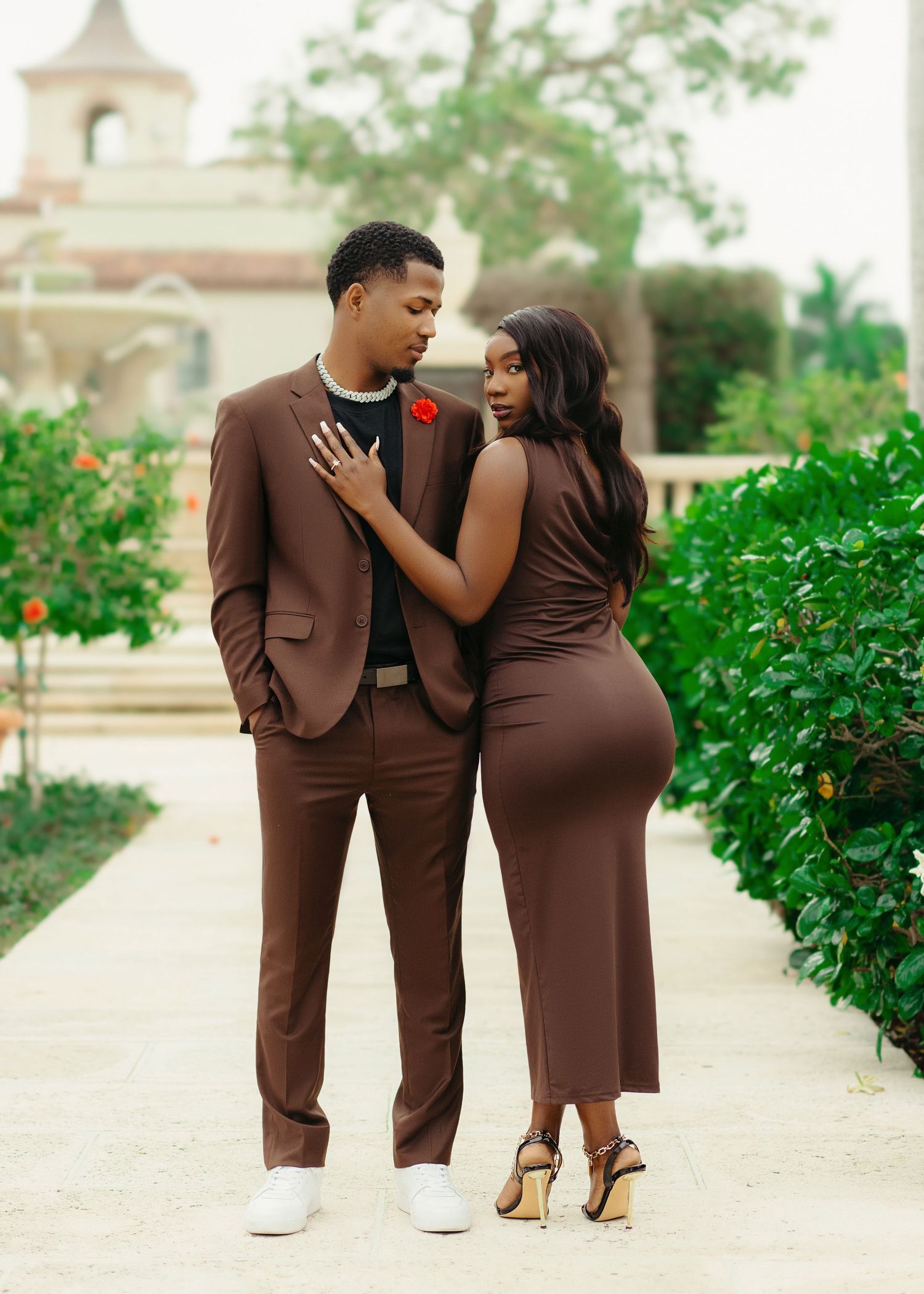 A couple in matching brown outfits pose together on a garden path with a church building visible in the background.
