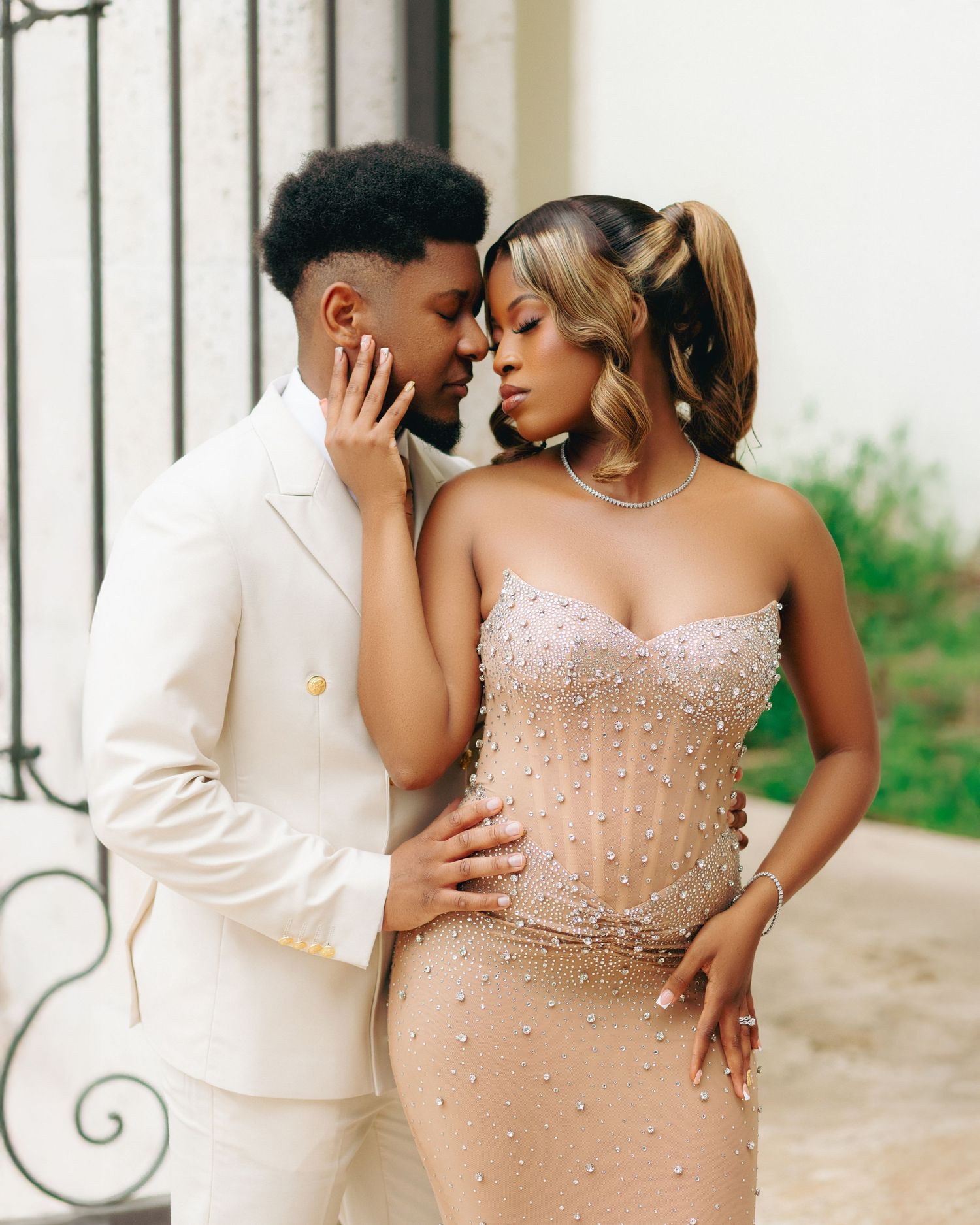 A romantic couple poses intimately against a wall, with one wearing a white suit and the other in a beaded gown.