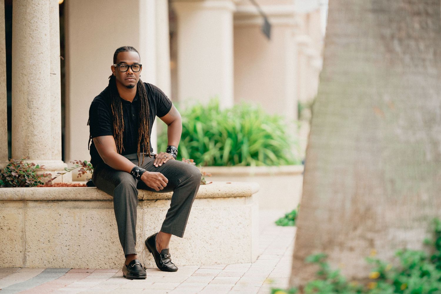 A person in a black shirt and jeans sits casually on a stone ledge near green plants and columns.