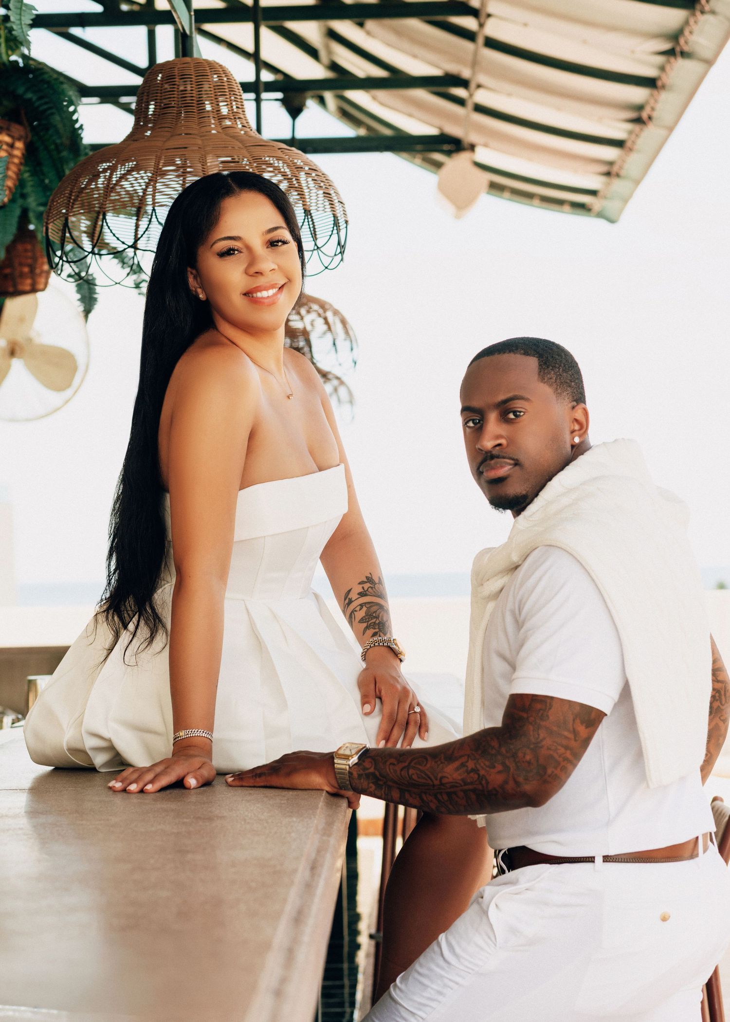 Couple in white outfits posing casually on covered patio with ocean view.