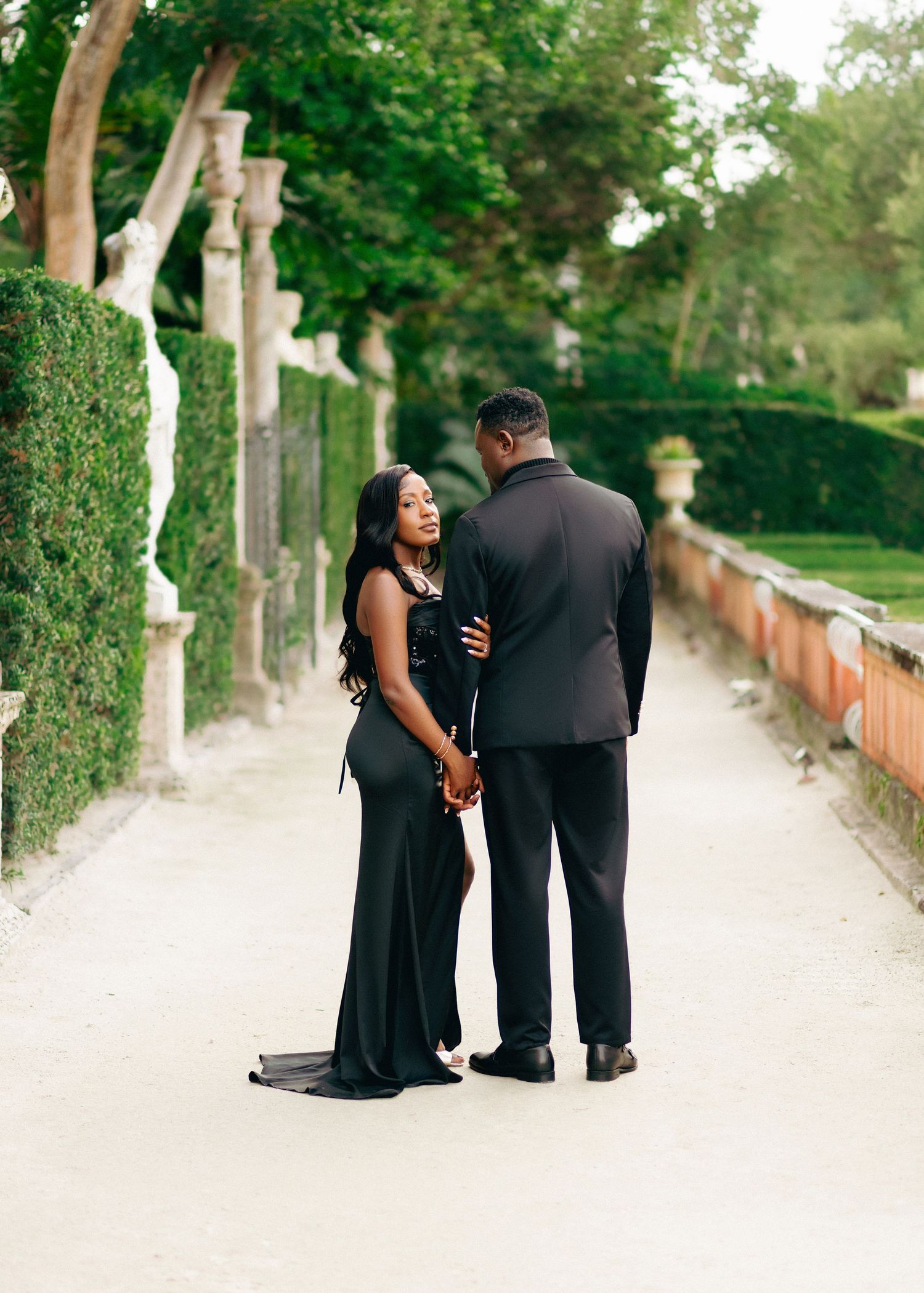 A couple in formal attire sharing a tender moment on a tree-lined garden path.