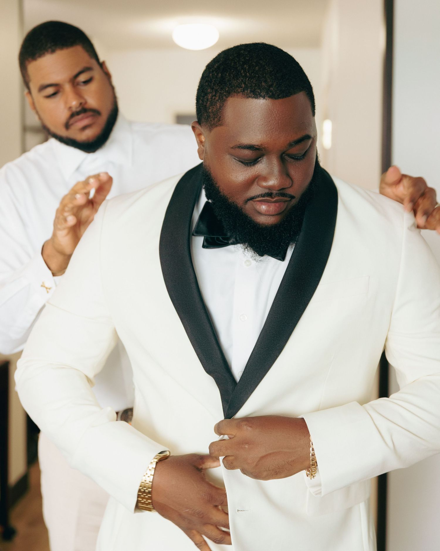 A groom in a white tuxedo with black shawl lapel adjusts his jacket.