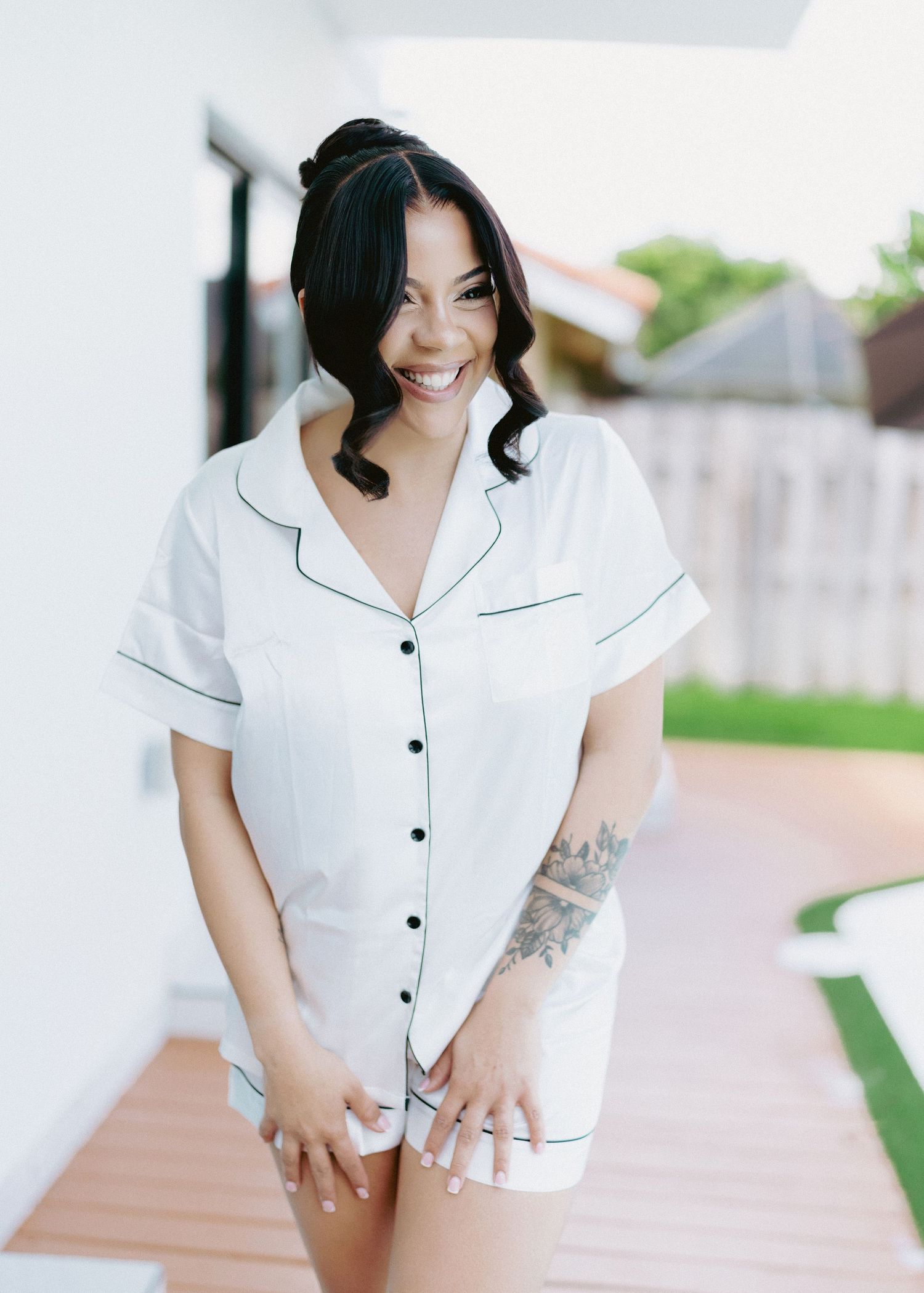 Smiling woman in white button-up pajama set with dark trim, standing on outdoor patio, showing floral arm tattoo.