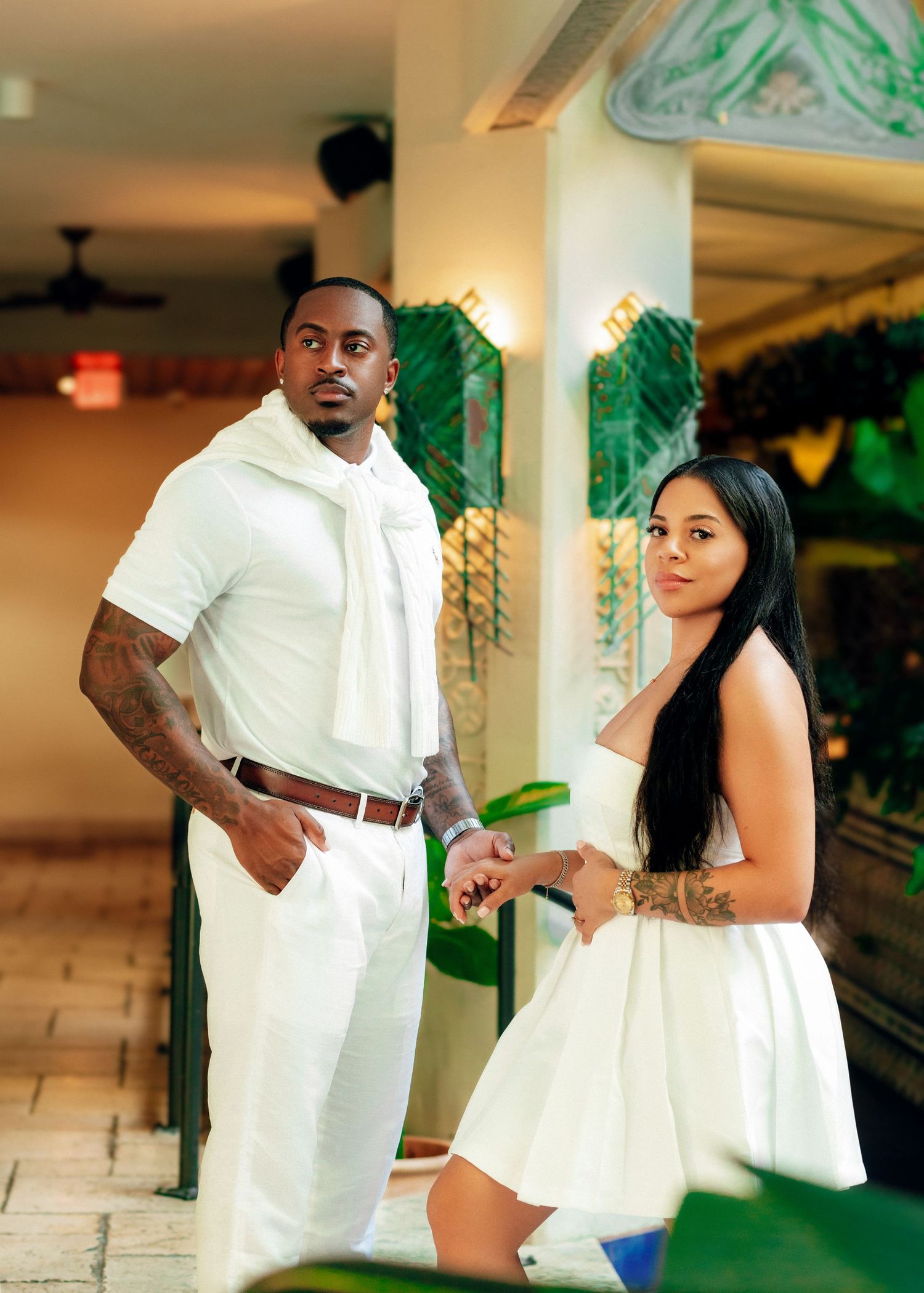 Elegant couple in white outfits poses together in an indoor setting with tropical plants.