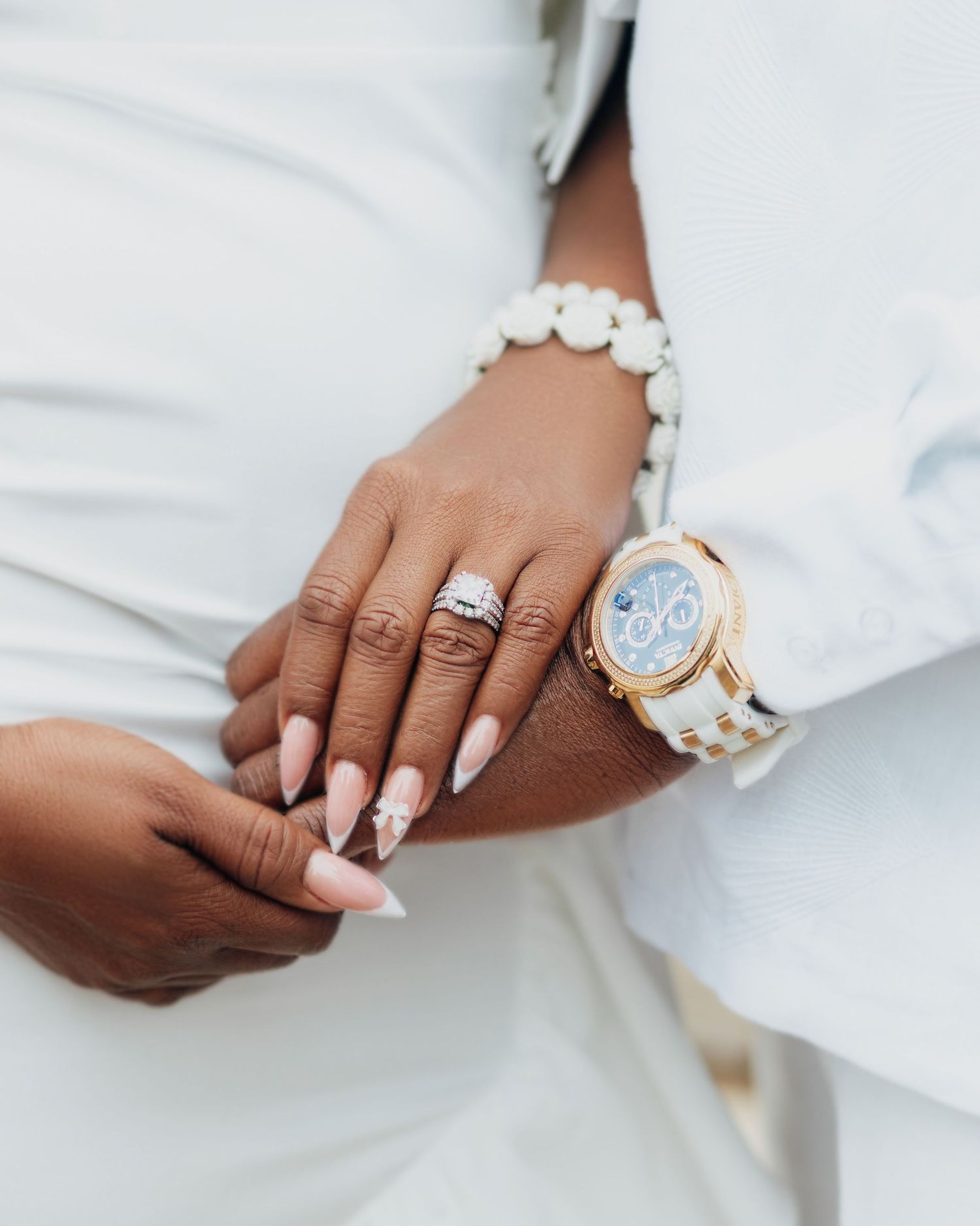 Close-up of hands showing engagement ring and wedding bands with white formal attire.
