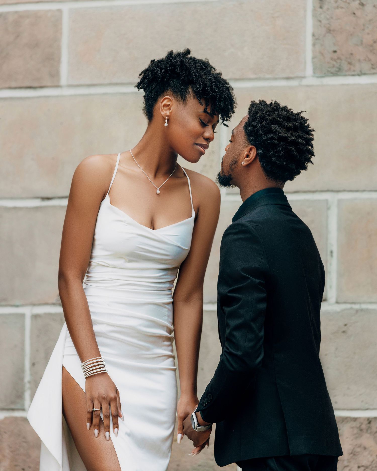 Romantic couple poses against stone wall in wedding attire.