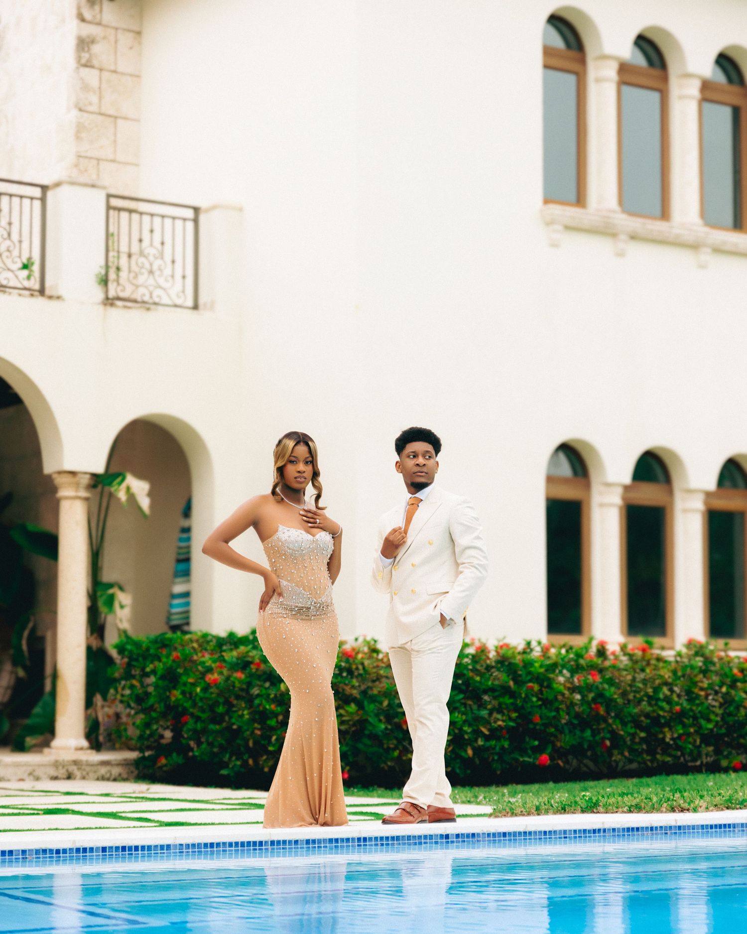 A couple in formal attire poses elegantly by a swimming pool at a Mediterranean-style venue with arched architecture.