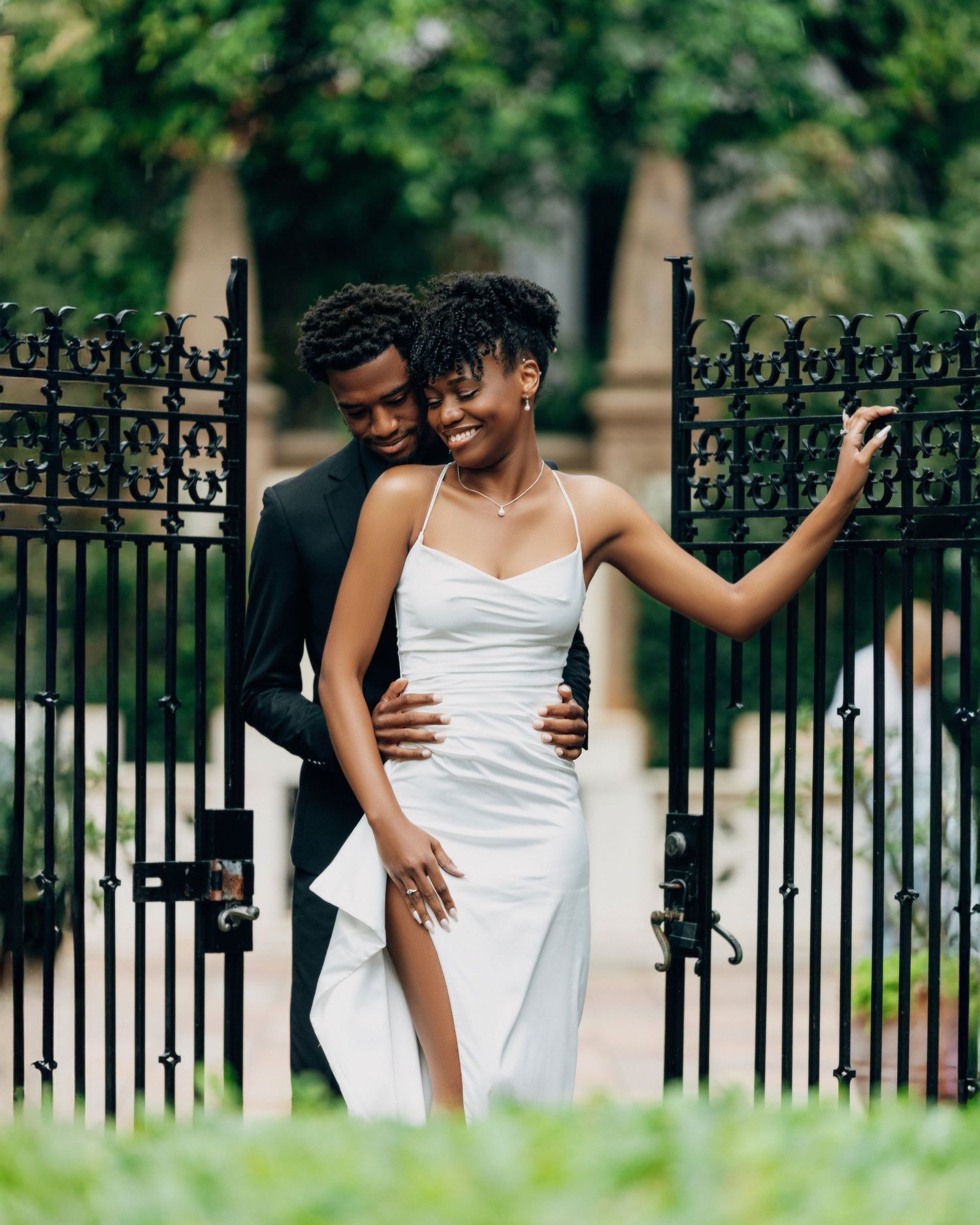 Elegant couple poses by decorative iron fence in garden setting.