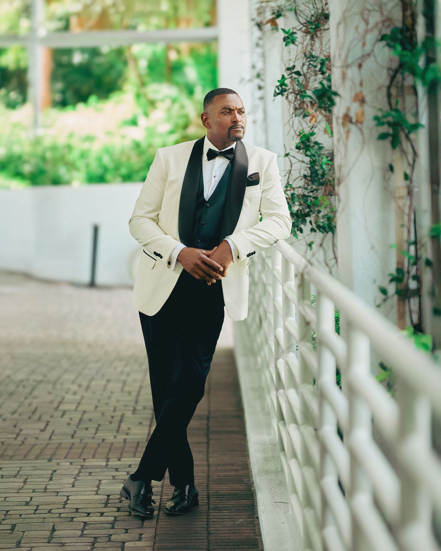 Groom in white tuxedo jacket poses along white railing in outdoor garden setting.