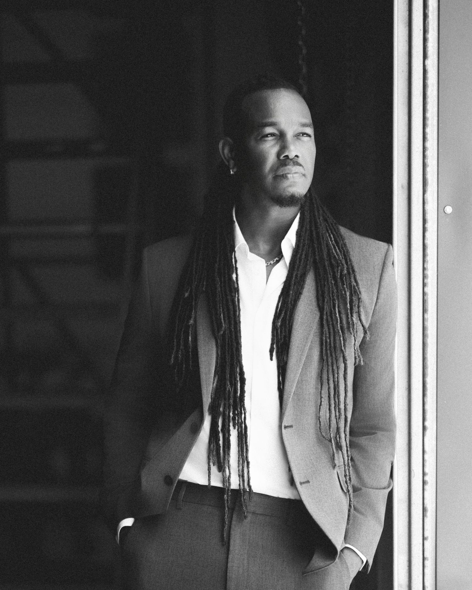 Black and white portrait of a man with long dreadlocks wearing a suit, gazing thoughtfully to the side.