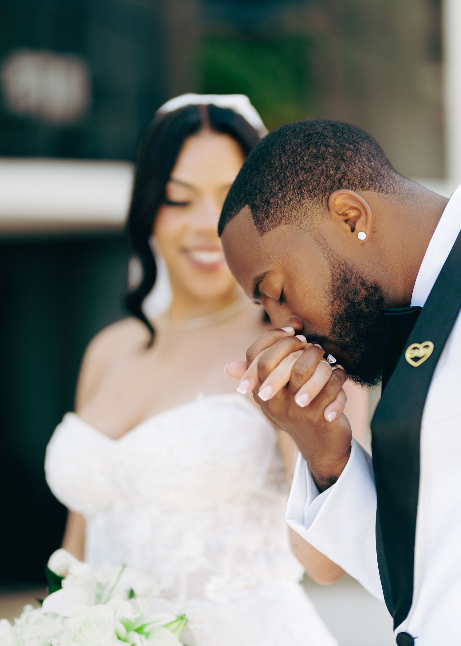Groom in white tuxedo kissing bride's hand during romantic outdoor wedding ceremony, bride smiling in strapless gown.