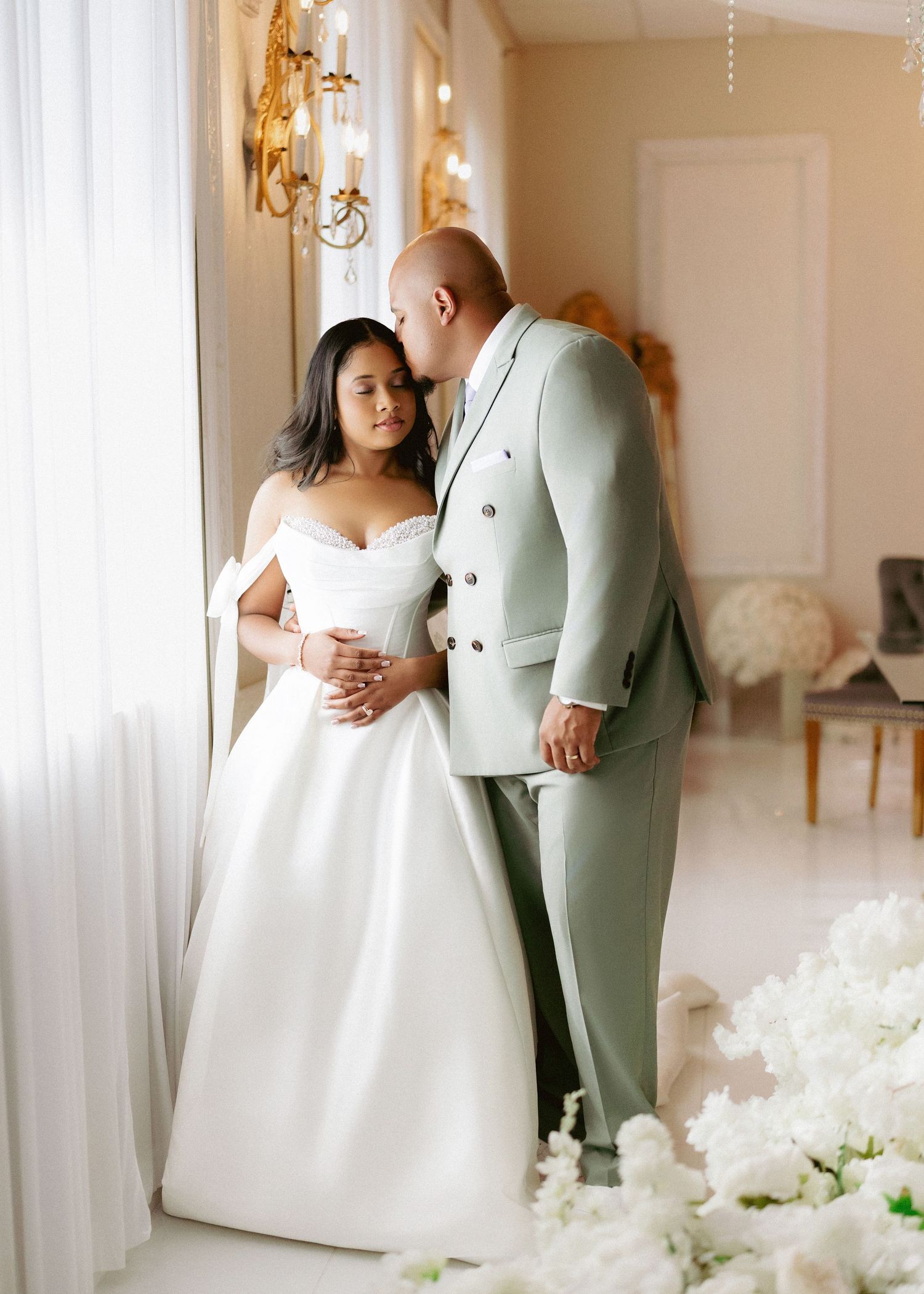 Bride in white off-shoulder gown and groom in sage green suit share intimate moment indoors.