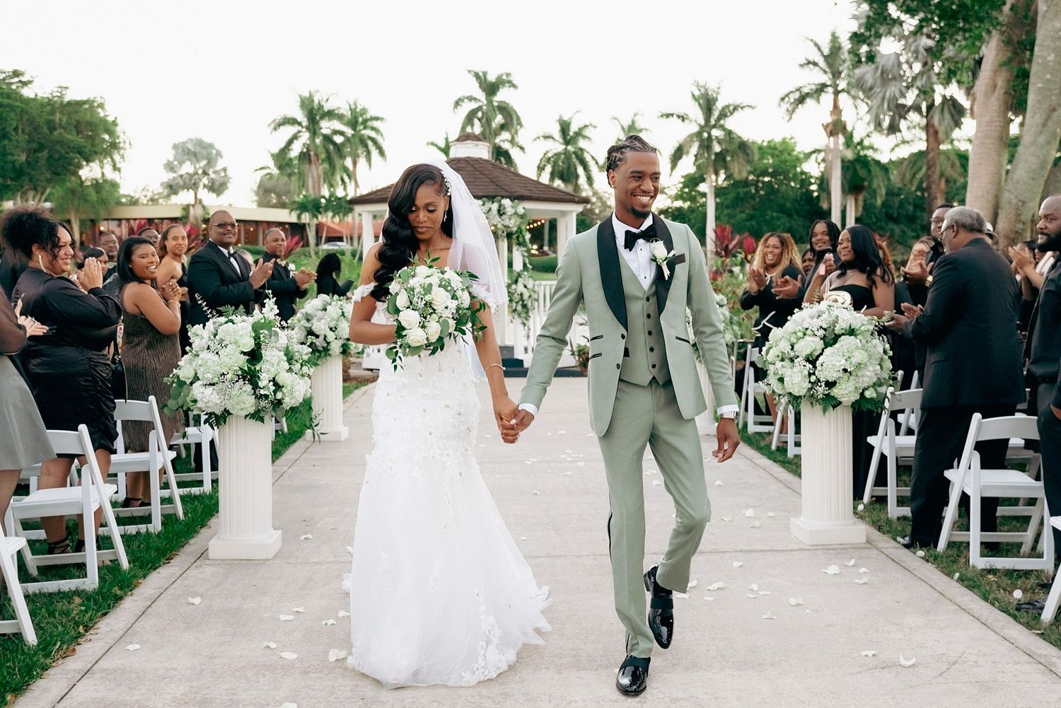 Wedding ceremony procession with couples walking down an outdoor aisle lined with white chairs and palm trees.