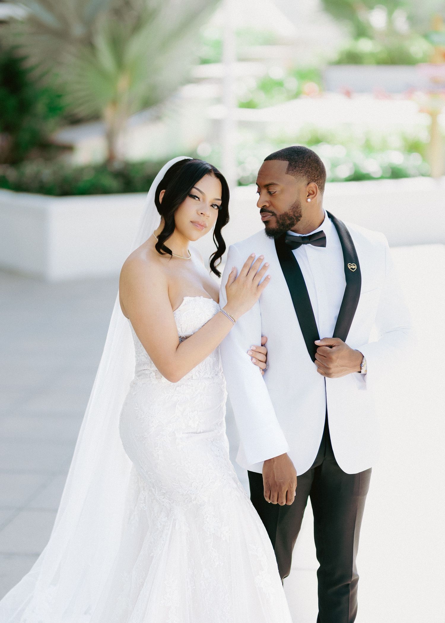 Bride in strapless lace mermaid gown with veil stands beside groom in white tuxedo jacket and black bow tie outdoors.