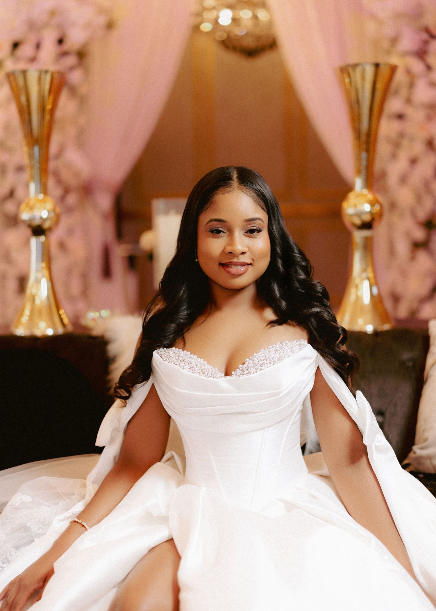 Bride in white off-shoulder ballgown with cape sits elegantly before pink floral backdrop.