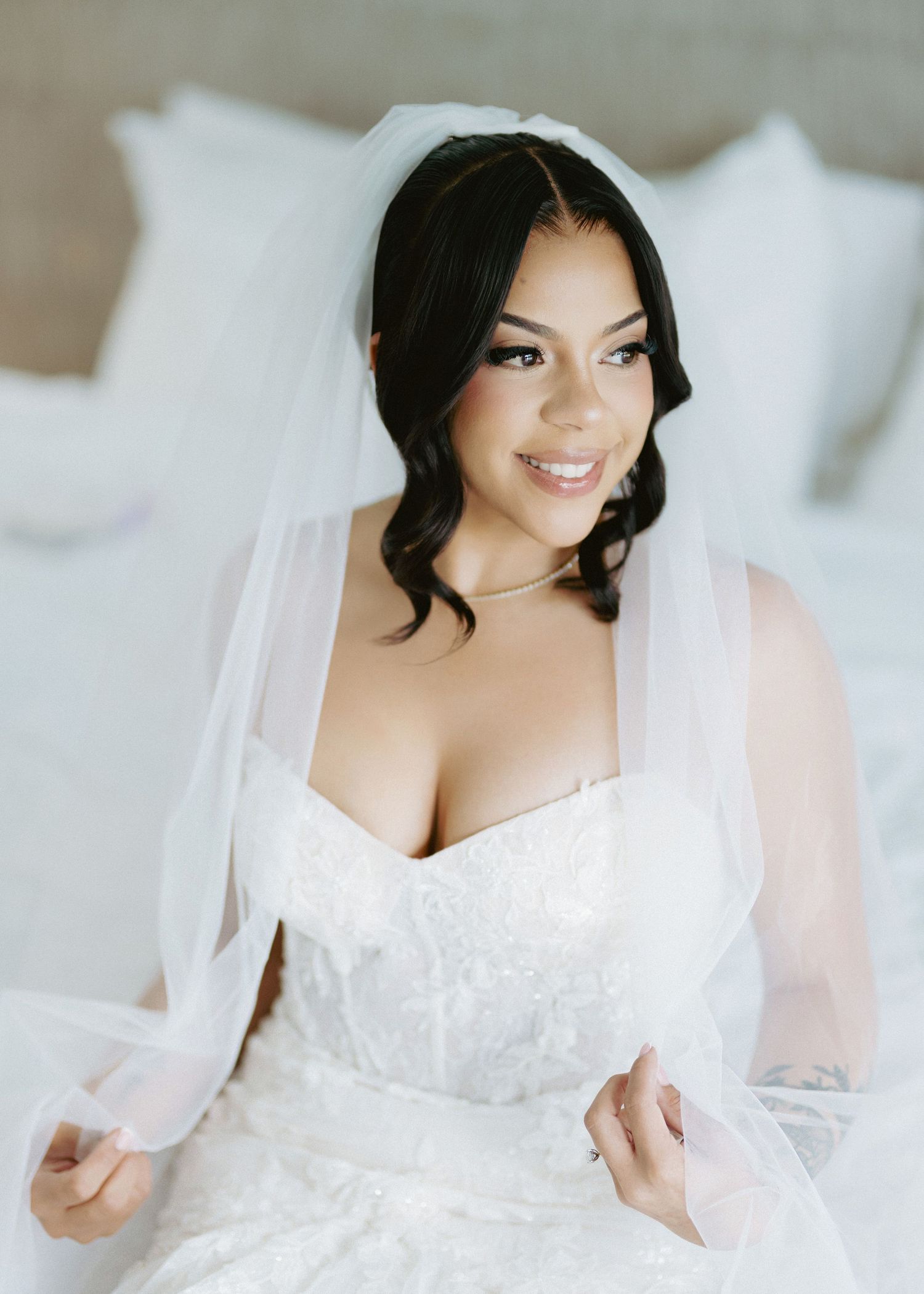 A beautiful bride in a white lace gown and veil sits on a bed, smiling softly on her wedding day.