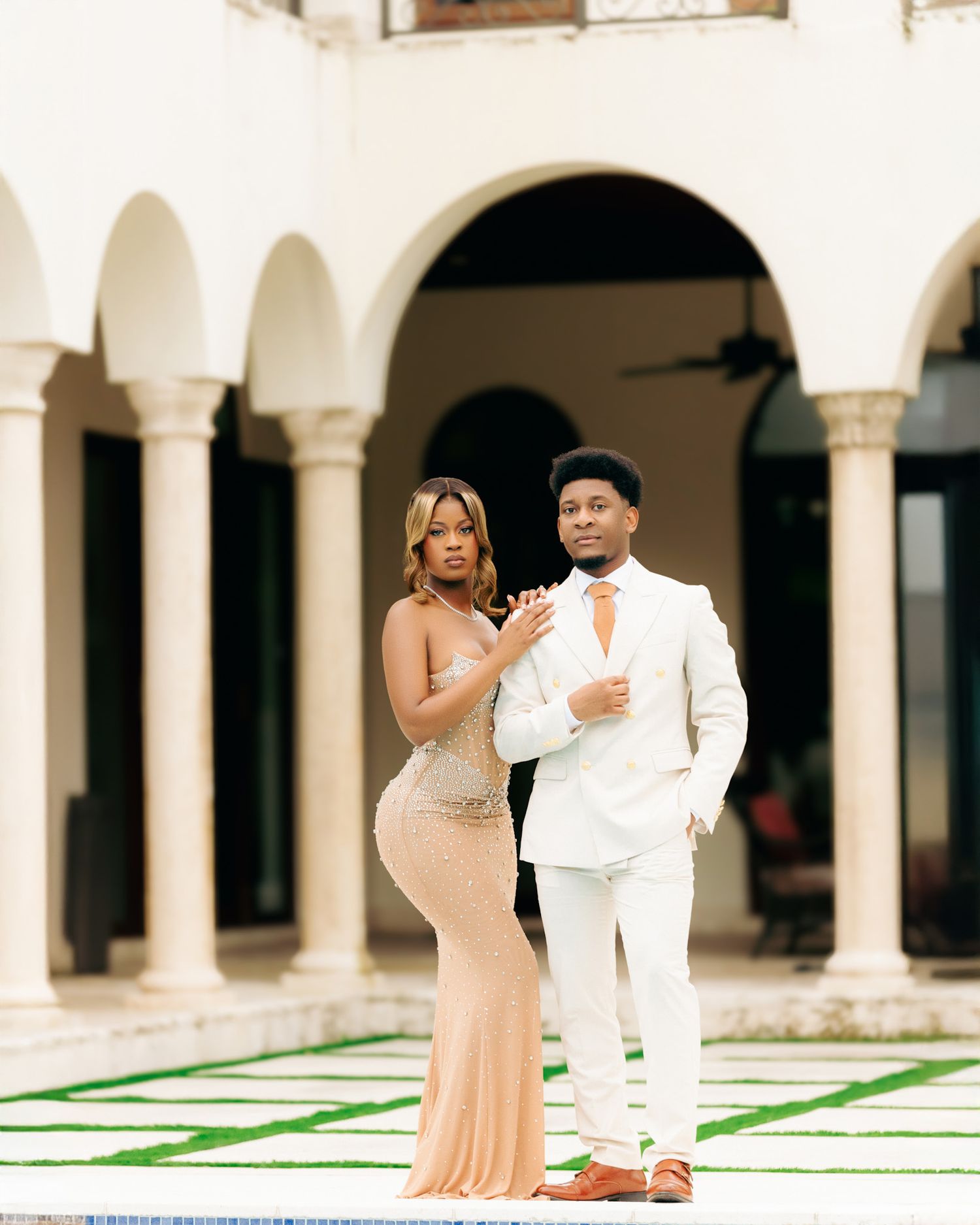 A couple in formal attire poses together under white arched columns in an elegant courtyard setting.