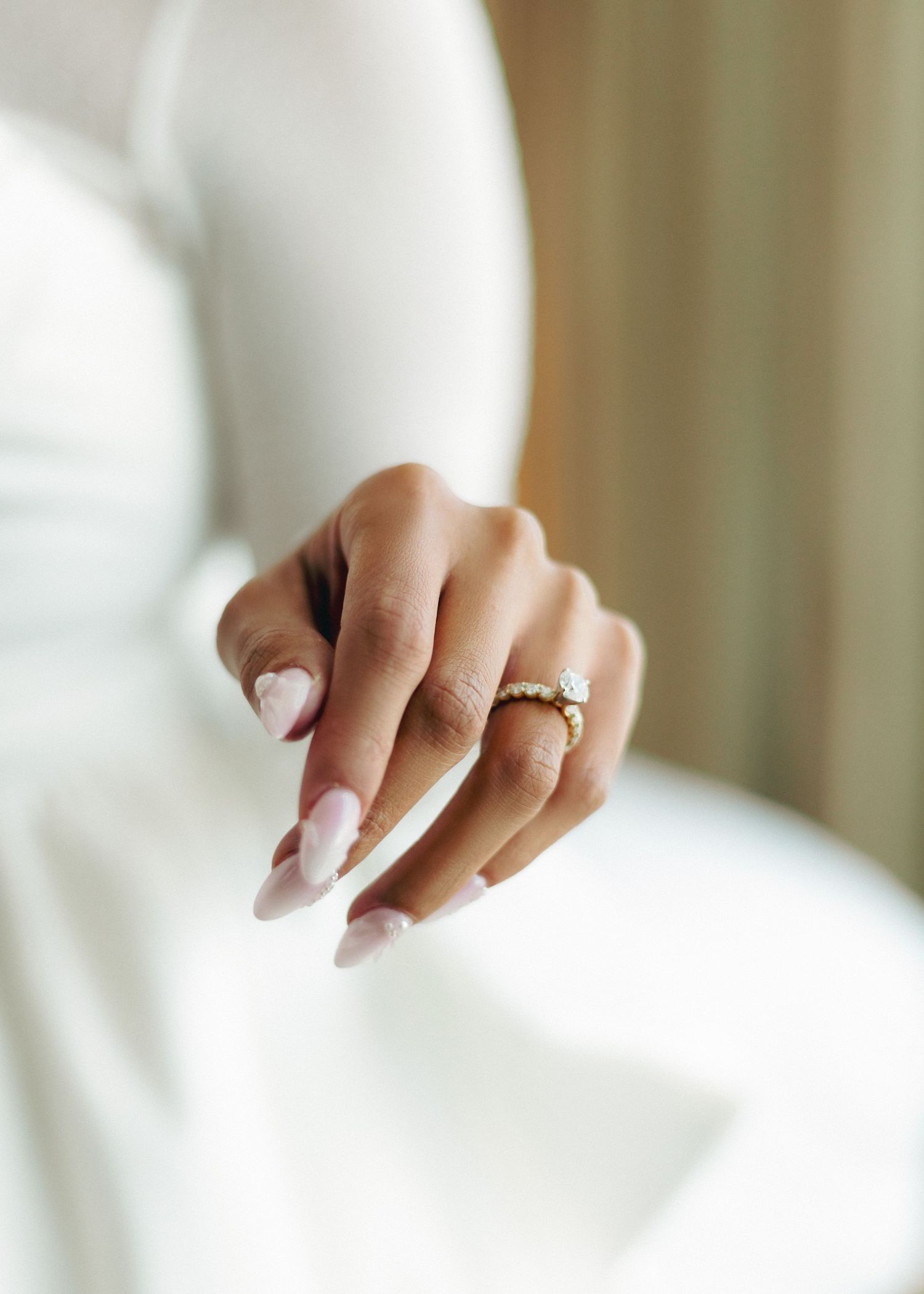 Close-up of a perfectly manicured hand with french tips wearing an elegant diamond engagement ring against white fabric.