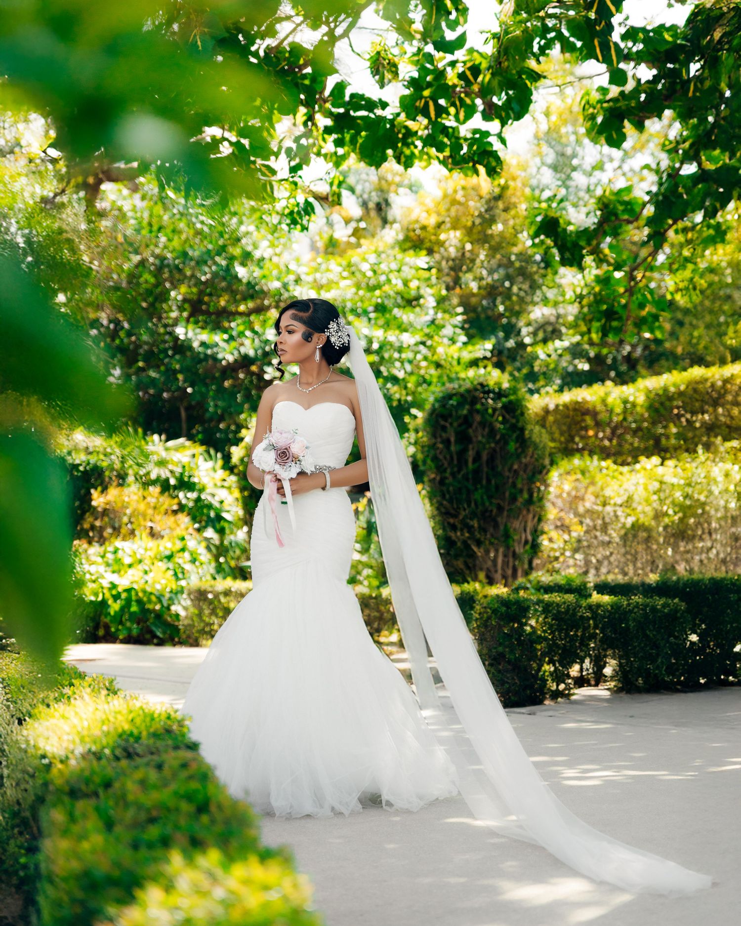 A bride in a flowing white gown and long veil stands in a sunlit garden setting surrounded by lush greenery.