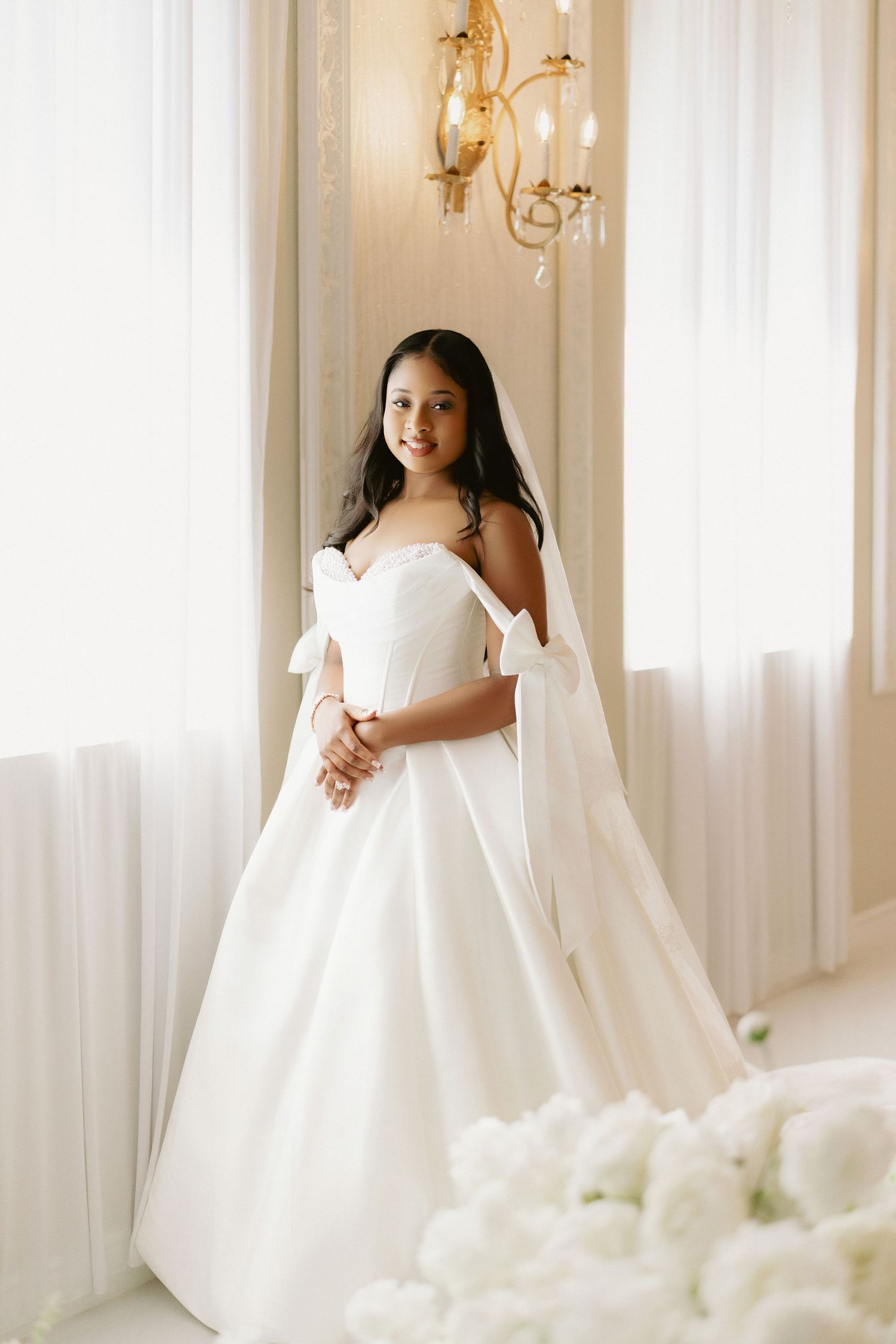 Elegant bride in strapless white ball gown with bow sleeves and veil, posing near bright windows with white flowers.