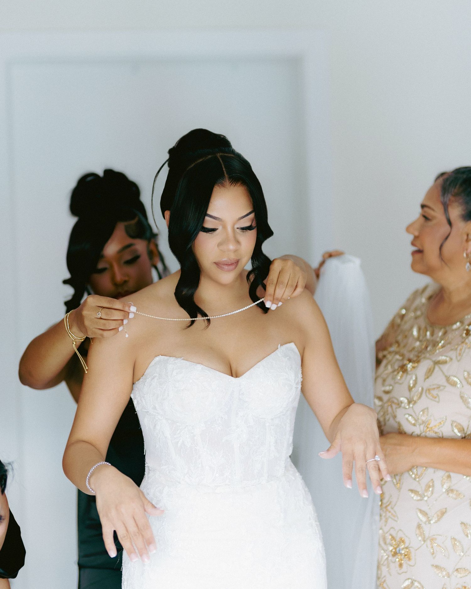 Bride in strapless lace wedding gown being helped with necklace by bridesmaids before ceremony.