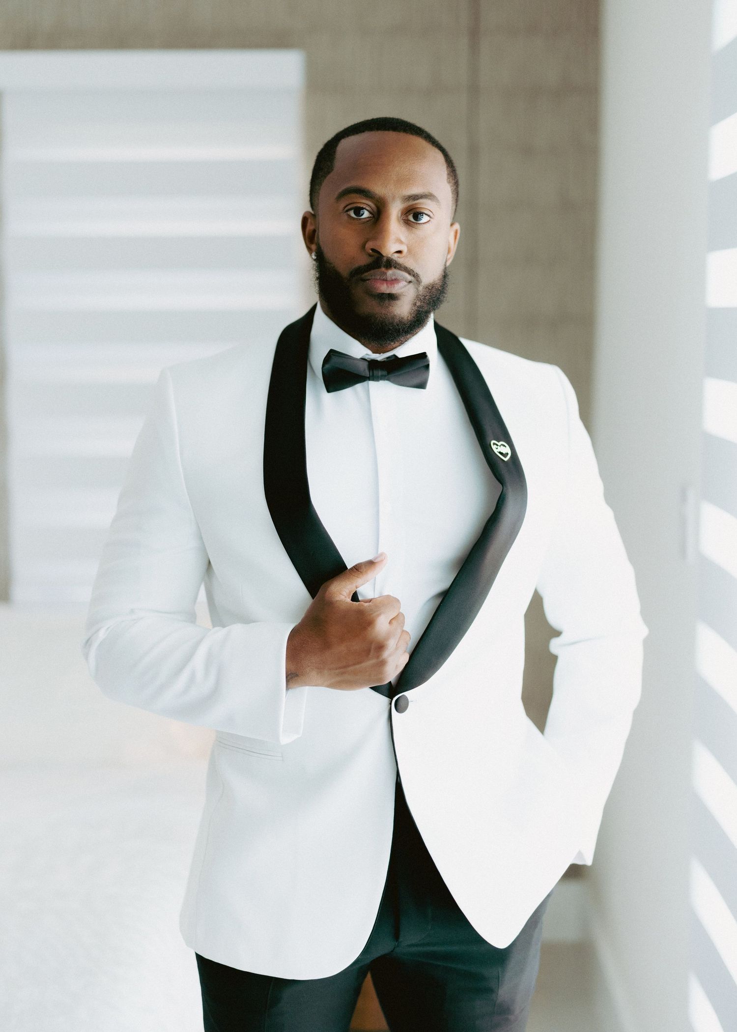 Groom in white tuxedo jacket with black shawl lapel and bow tie posing confidently indoors.