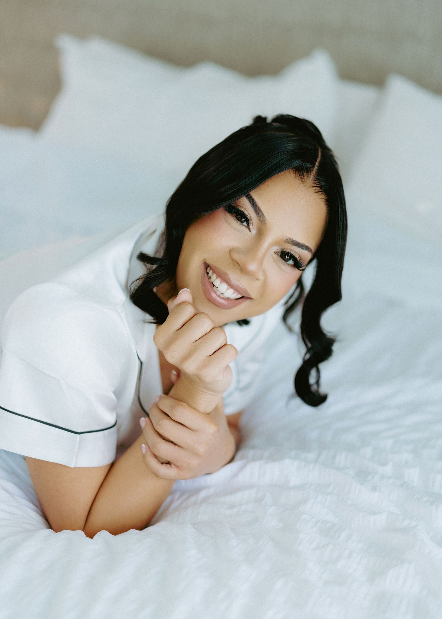 Smiling young woman with dark hair lying on white bedding wearing a white top.