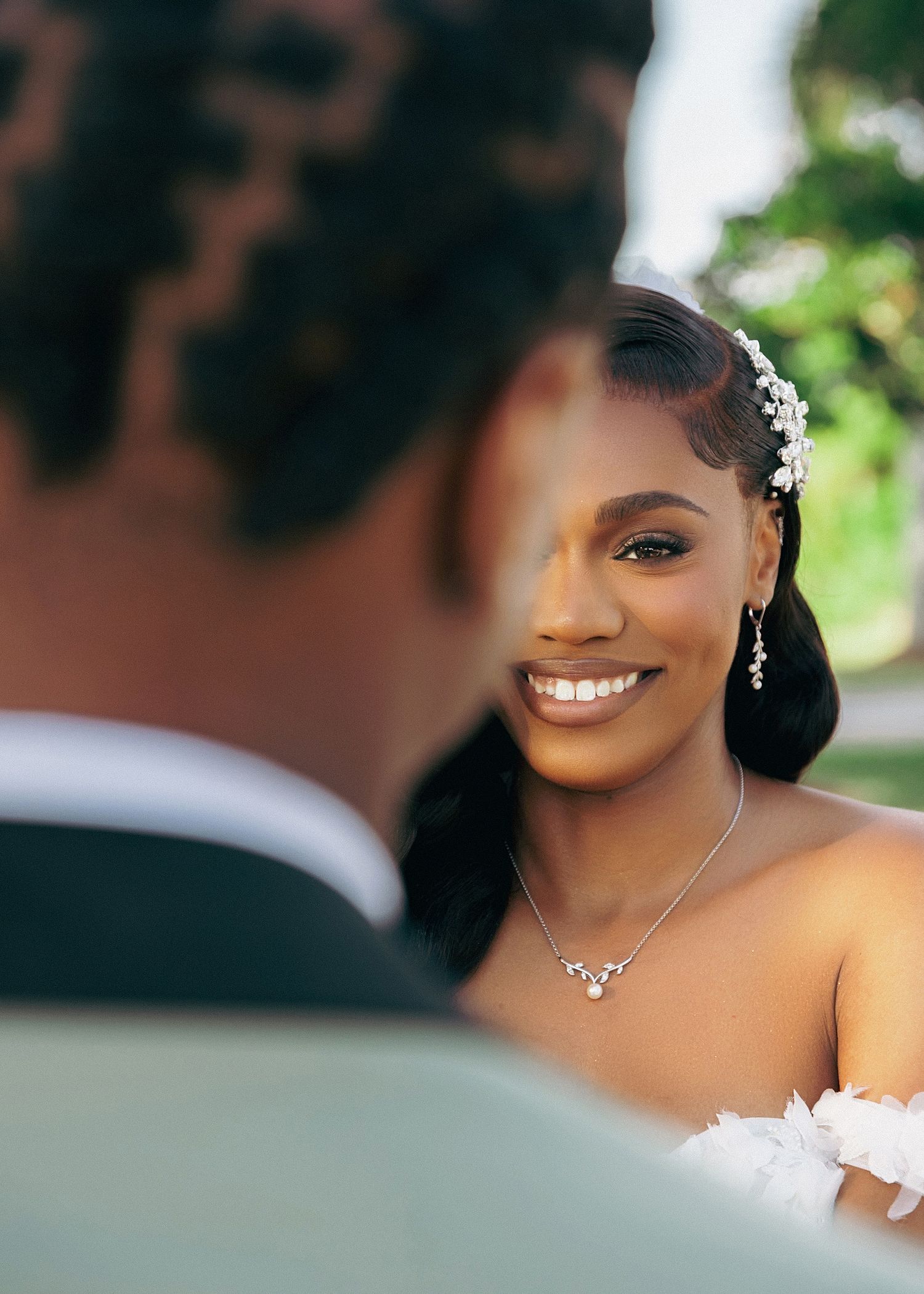 A close-up wedding portrait shows a bride wearing delicate jewelry and an off-shoulder white dress in a garden setting.