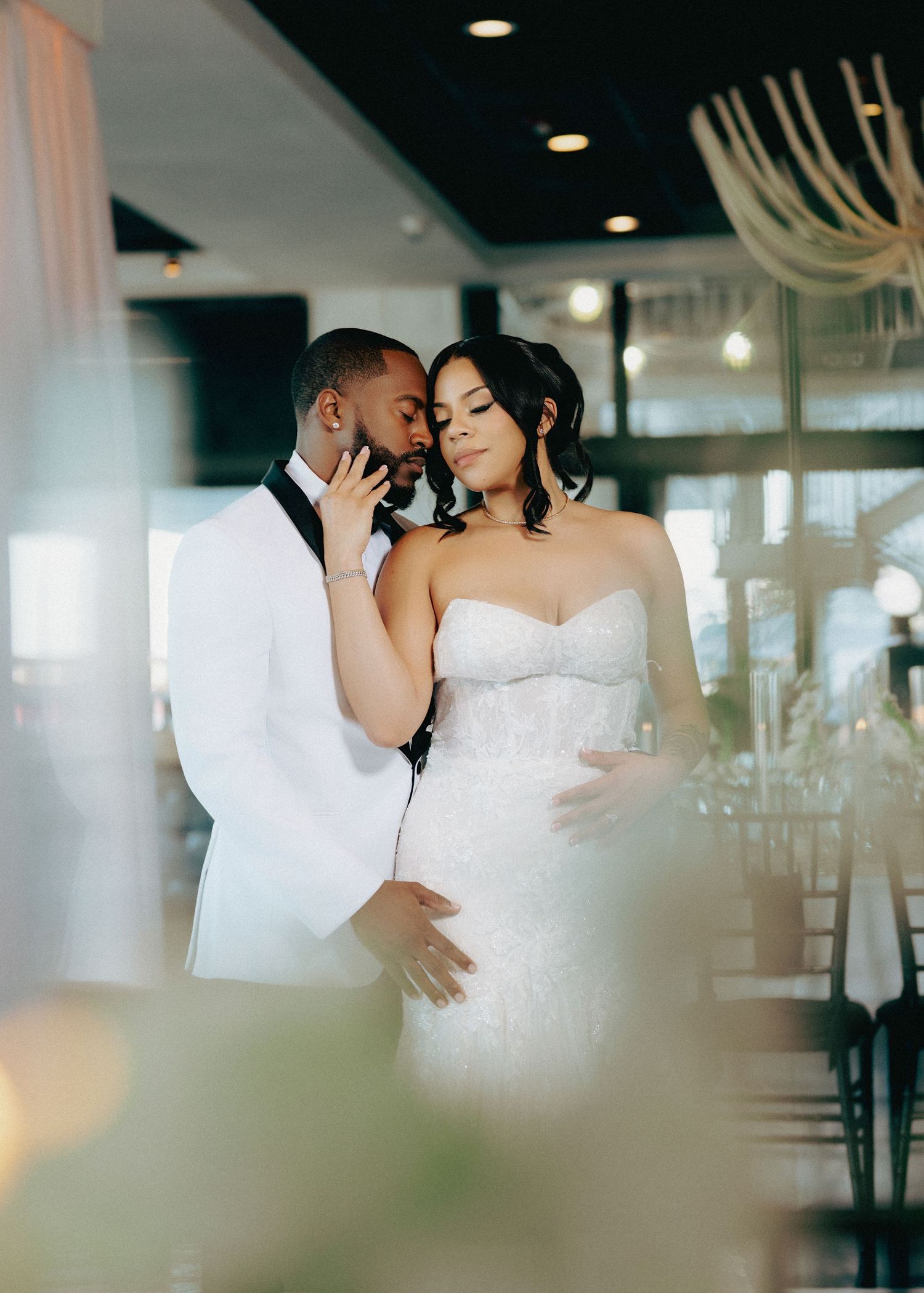 Elegant bride and groom embracing in a stunning wedding venue, bride in beaded strapless gown, groom in white tuxedo.