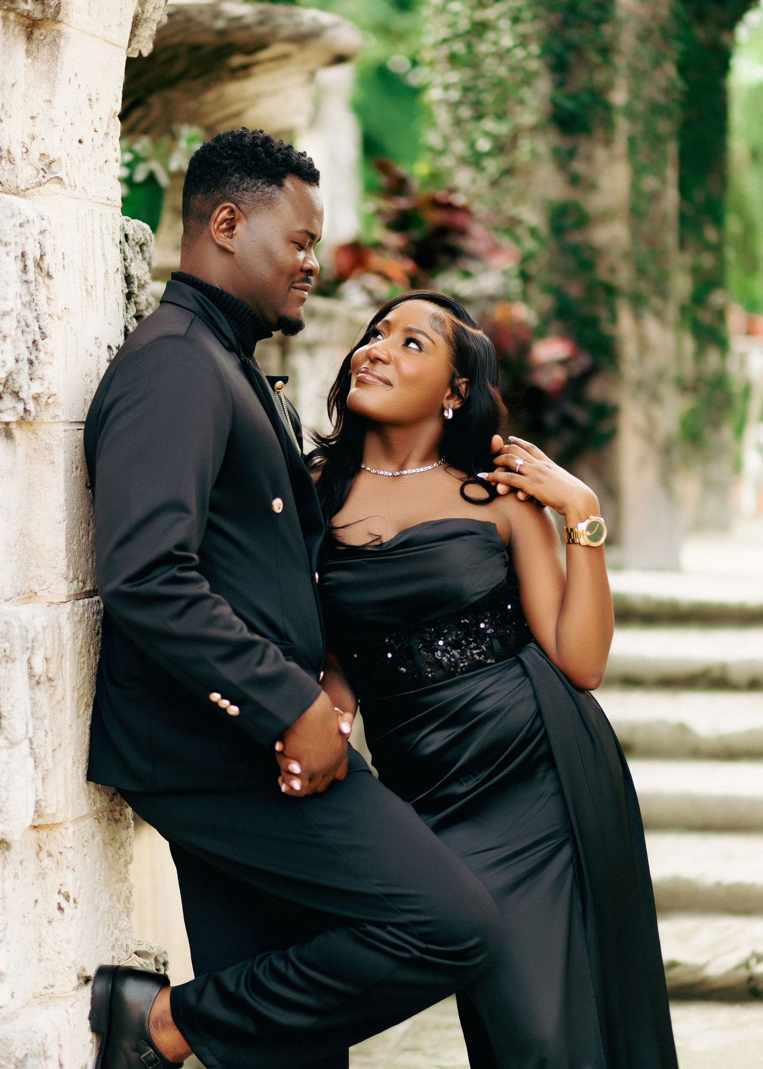 A romantic couple in elegant black formal wear pose together against a stone pillar in a garden setting.